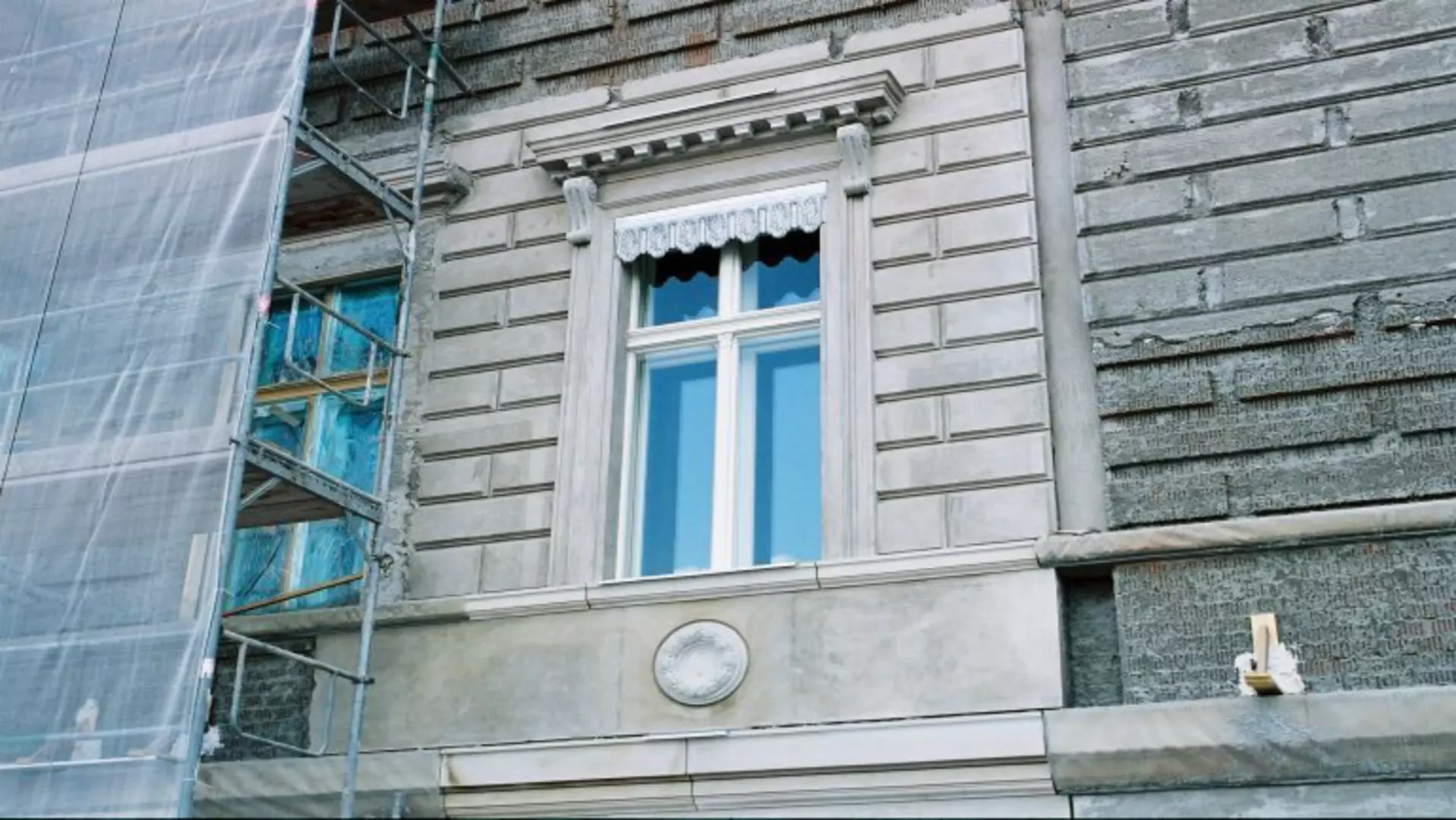 Fassadenausschnitt mit eingerüsteter Wand, klassizistischem Fenster mit Zierrahmung und erneuerter Oberfläche während Bauarbeiten.