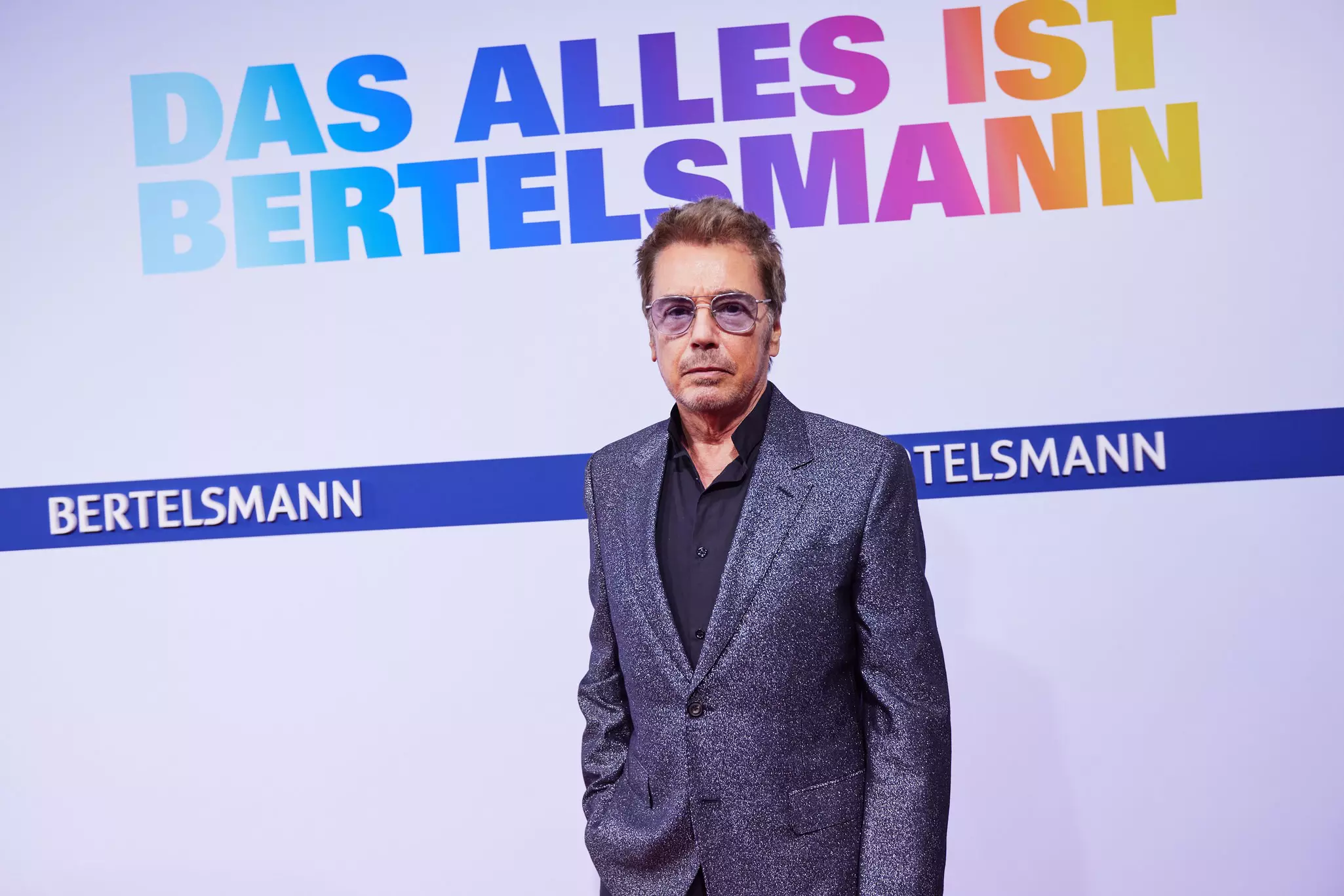 Jean-Michel Jarre mit Sonnenbrille und glitzerndem Sakko vor Bertelsmann-Wand mit Slogan „DAS ALLES IST BERTELSMANN“
