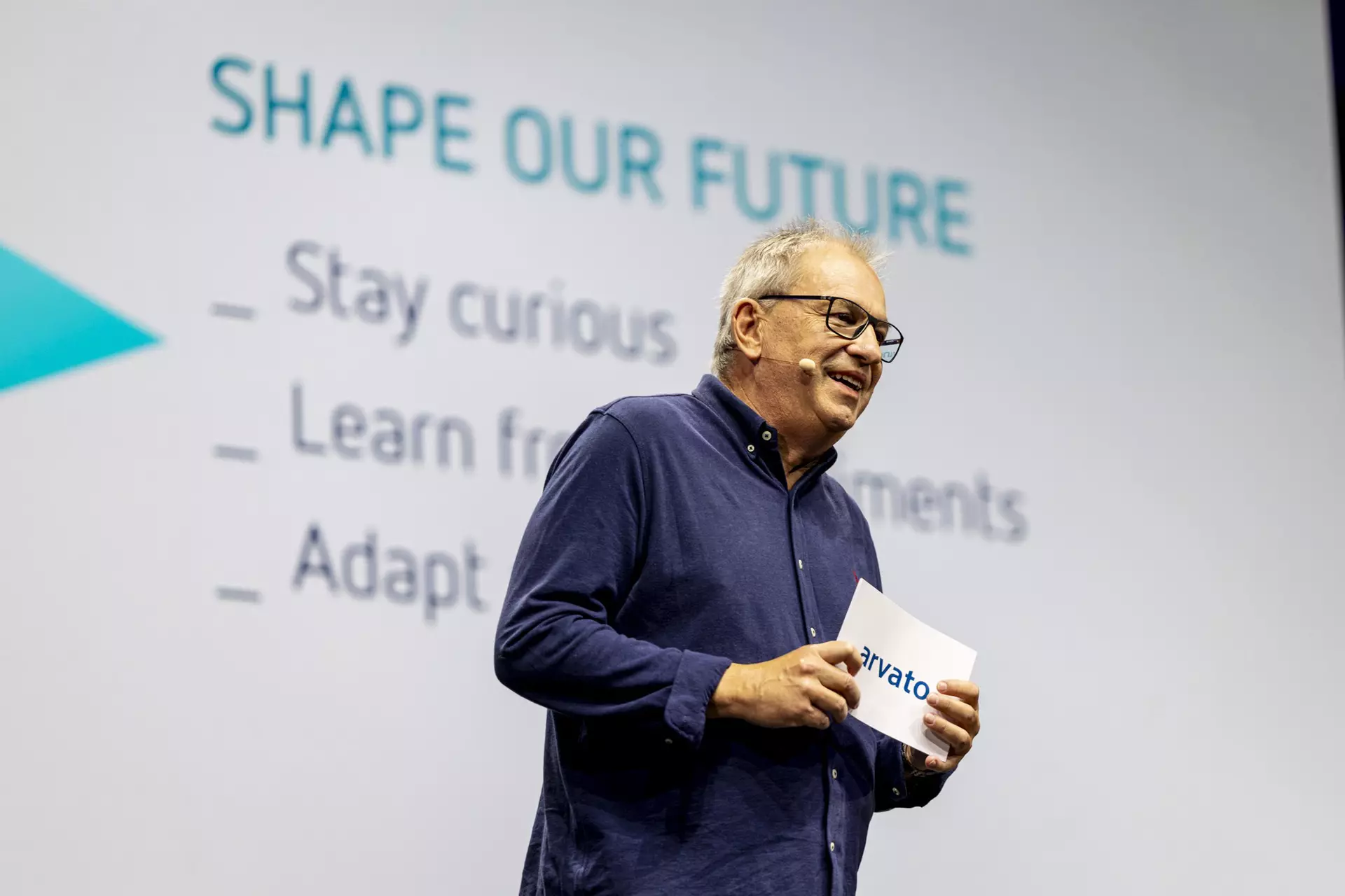 Person mit Brille hält Karte auf Bühne vor Folie mit Titel „SHAPE OUR FUTURE“.