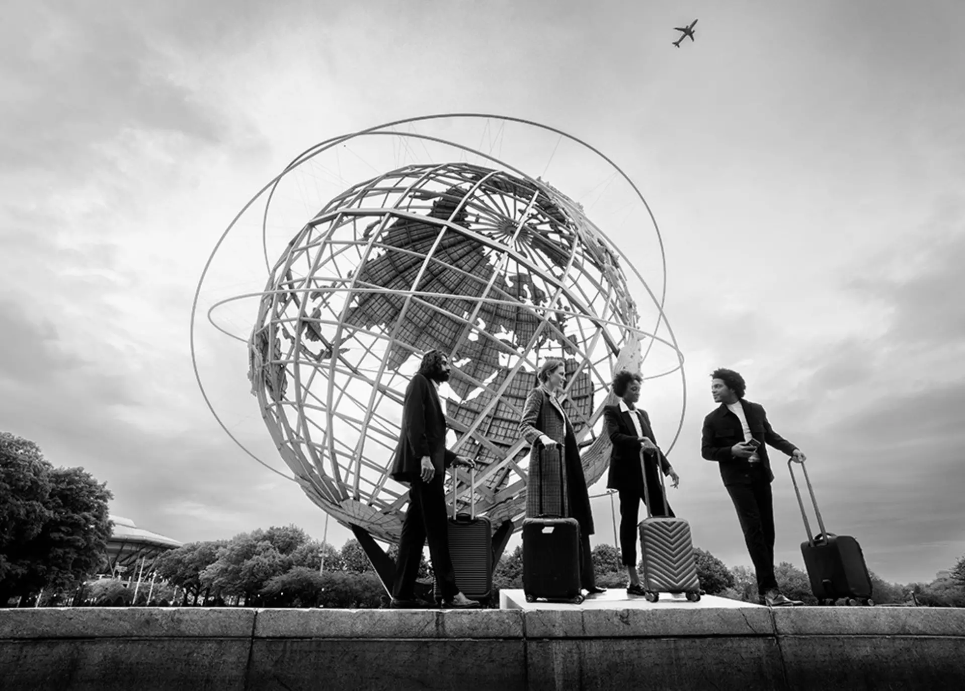 Vier Geschäftsreisende mit Rollkoffern stehen vor der Unisphere in New York City; über ihnen fliegt Flugzeug am bewölkten Himmel.