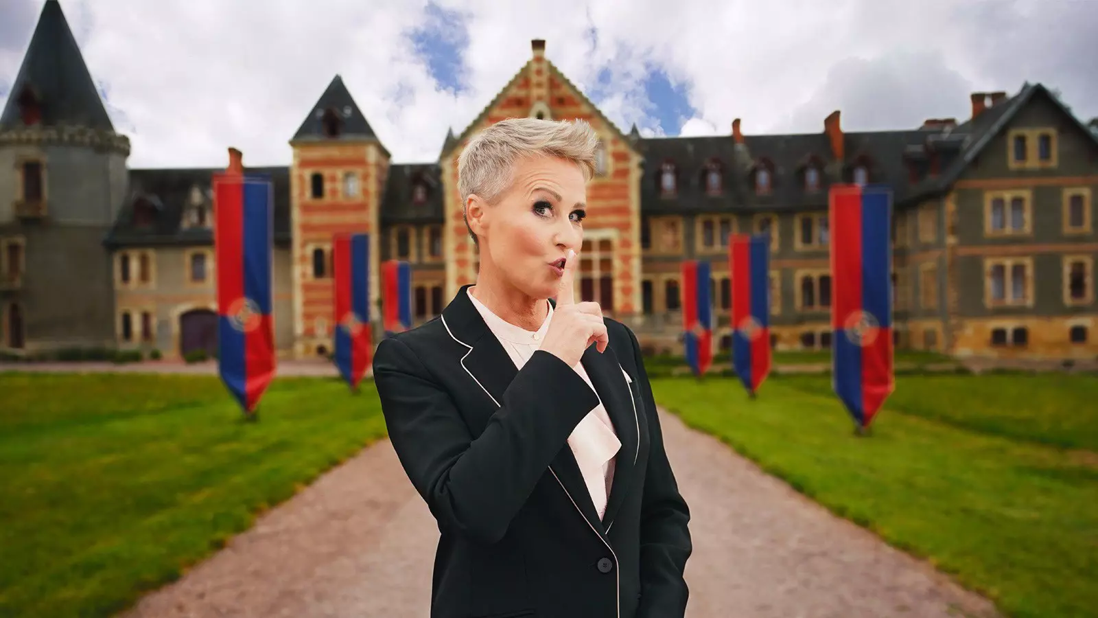 The presenter Sonja Zietlow stands in front of a castle-like building with red and blue banners and puts her finger to her lips.