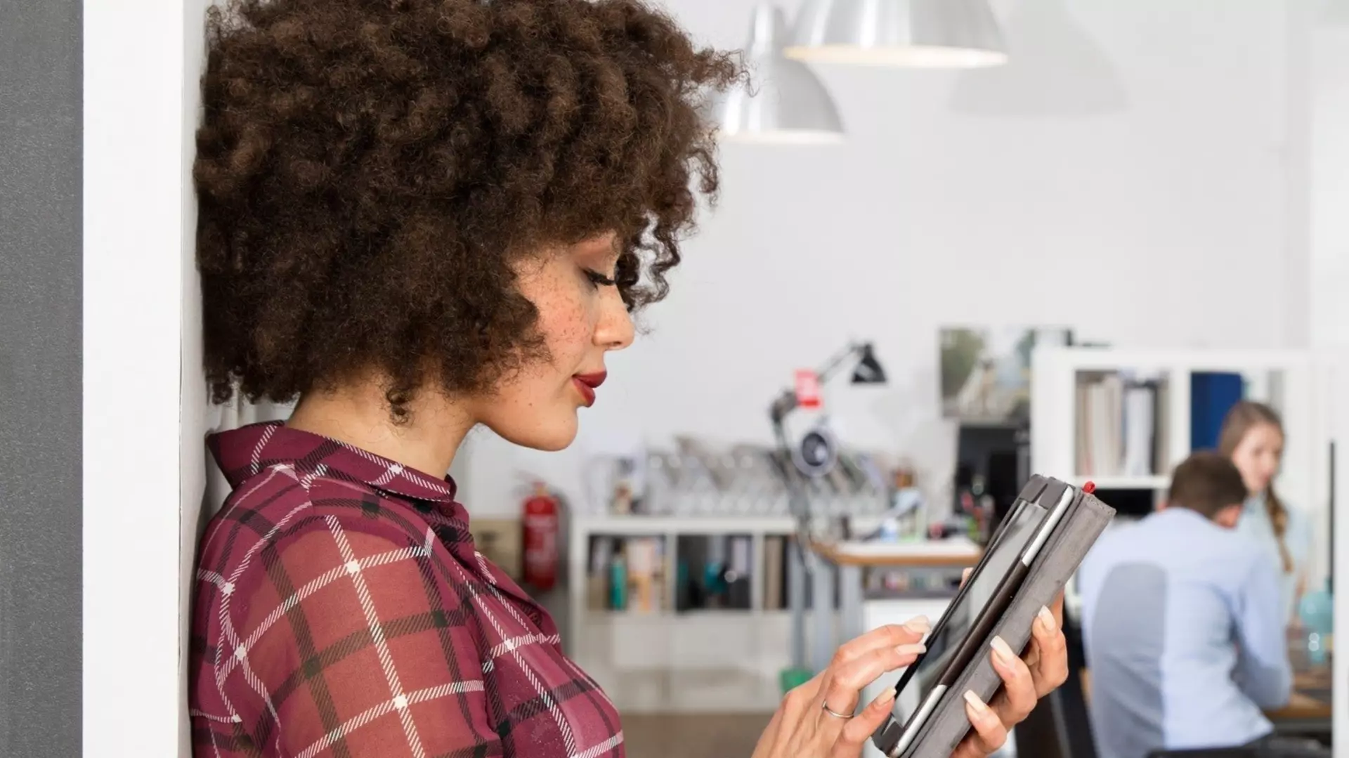 A woman with curly hair is leaning against a wall and reading on a tablet; in the background, a modern office with people working can be seen out of focus.