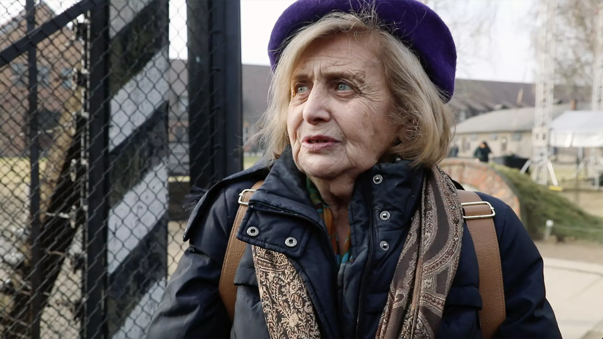 Person wearing a winter coat and scarf stands in front of a fence and buildings in a historic outdoor site.