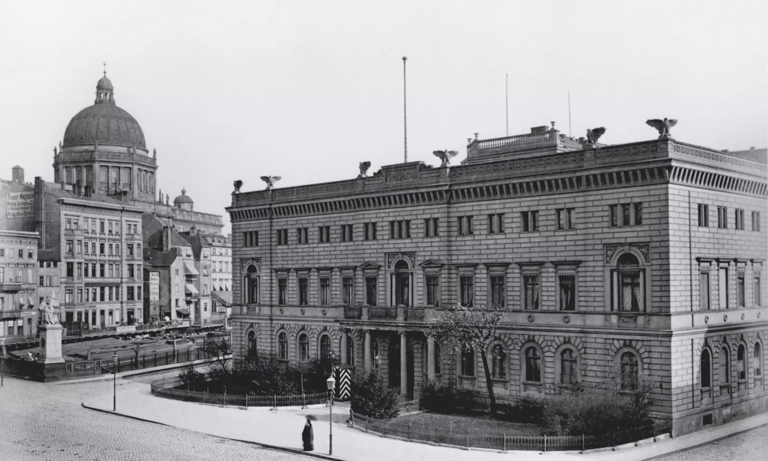 Historische Fotografie mit zwei großen klassizistischen Gebäuden an einem städtischen Platz, links ein Kuppelbau, rechts dreigeschossiges Palais mit Balkon und kleinem Vorgarten.