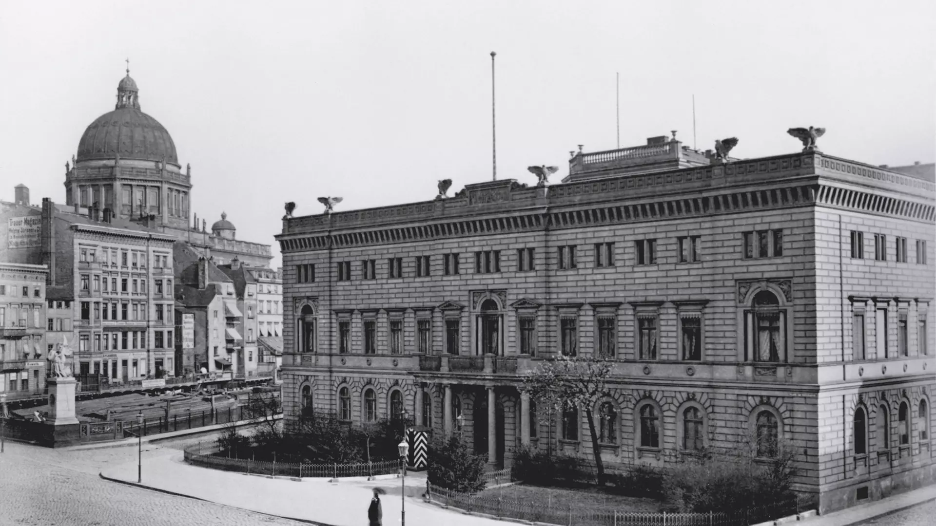 Historische Fotografie mit zwei großen klassizistischen Gebäuden an einem städtischen Platz, links ein Kuppelbau, rechts dreigeschossiges Palais mit Balkon und kleinem Vorgarten.