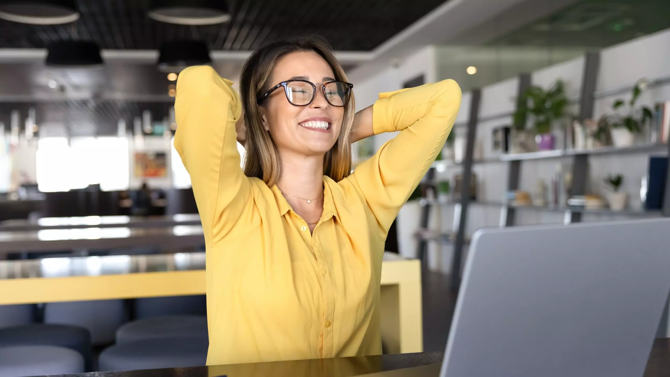 Person in gelber Bluse sitzt vor einem Laptop in einem modernen Büro und lehnt sich entspannt mit verschränkten Armen zurück.