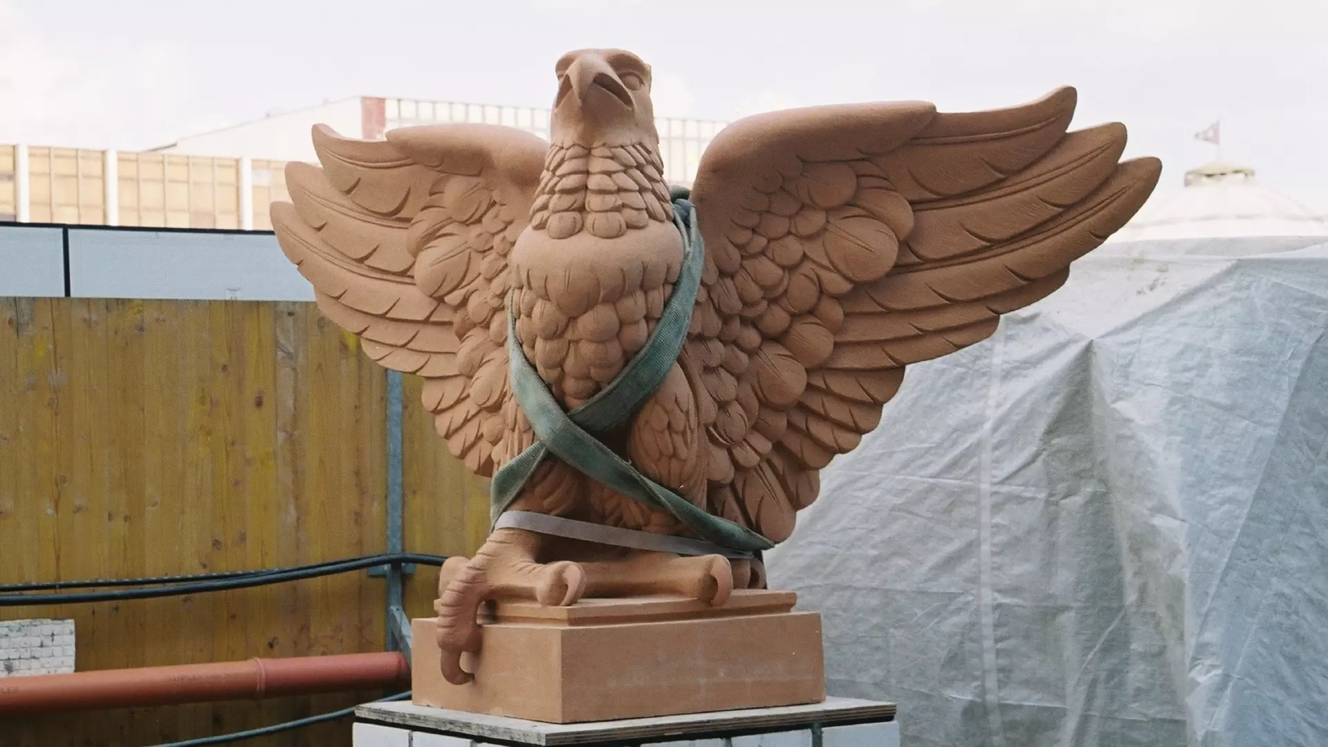 Skulptur eines Adlers mit ausgebreiteten Flügeln, mit Schutzband umwickelt, auf Sockel in Werkstattumgebung im Freien.