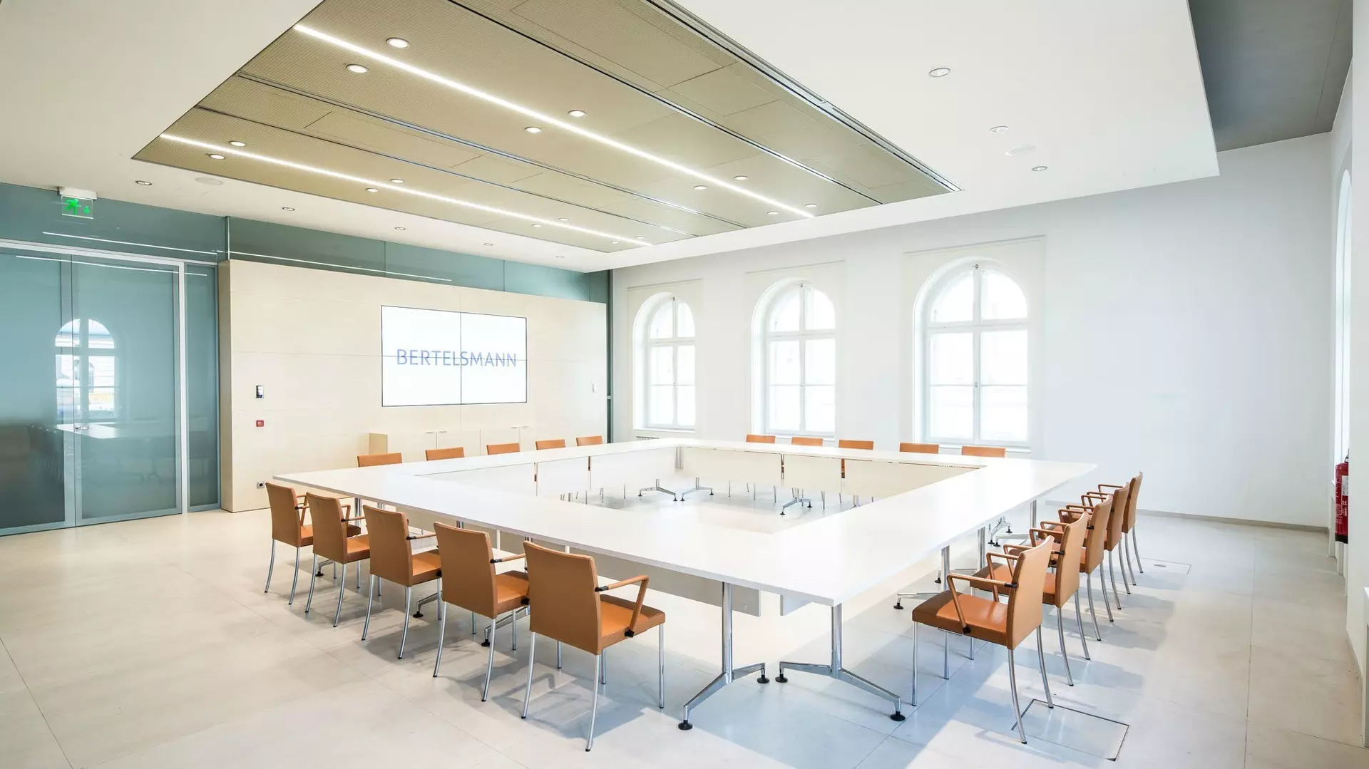 Moderner Konferenzraum mit zum Quadrat angeordneten Tischen, hellbraunen Stühlen, LED-Bildschirm und hohen Rundbogenfenstern.