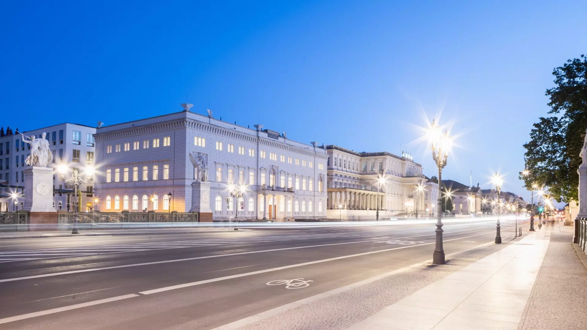 Steinerne Brücke mit Statuen bei Dämmerung, beleuchtete Gebäude im Hintergrund, ruhiger Fluss spiegelt Licht