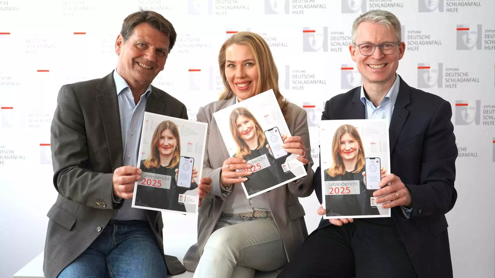 Three people present the 2025 annual report of the German Stroke Foundation, holding copies toward the camera.