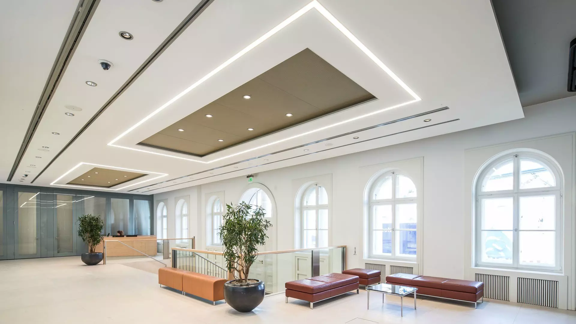 Großzügiges Foyer mit Sitzbänken, Glastischen, Pflanzen, Rundbogenfenstern und moderner Deckenbeleuchtung.