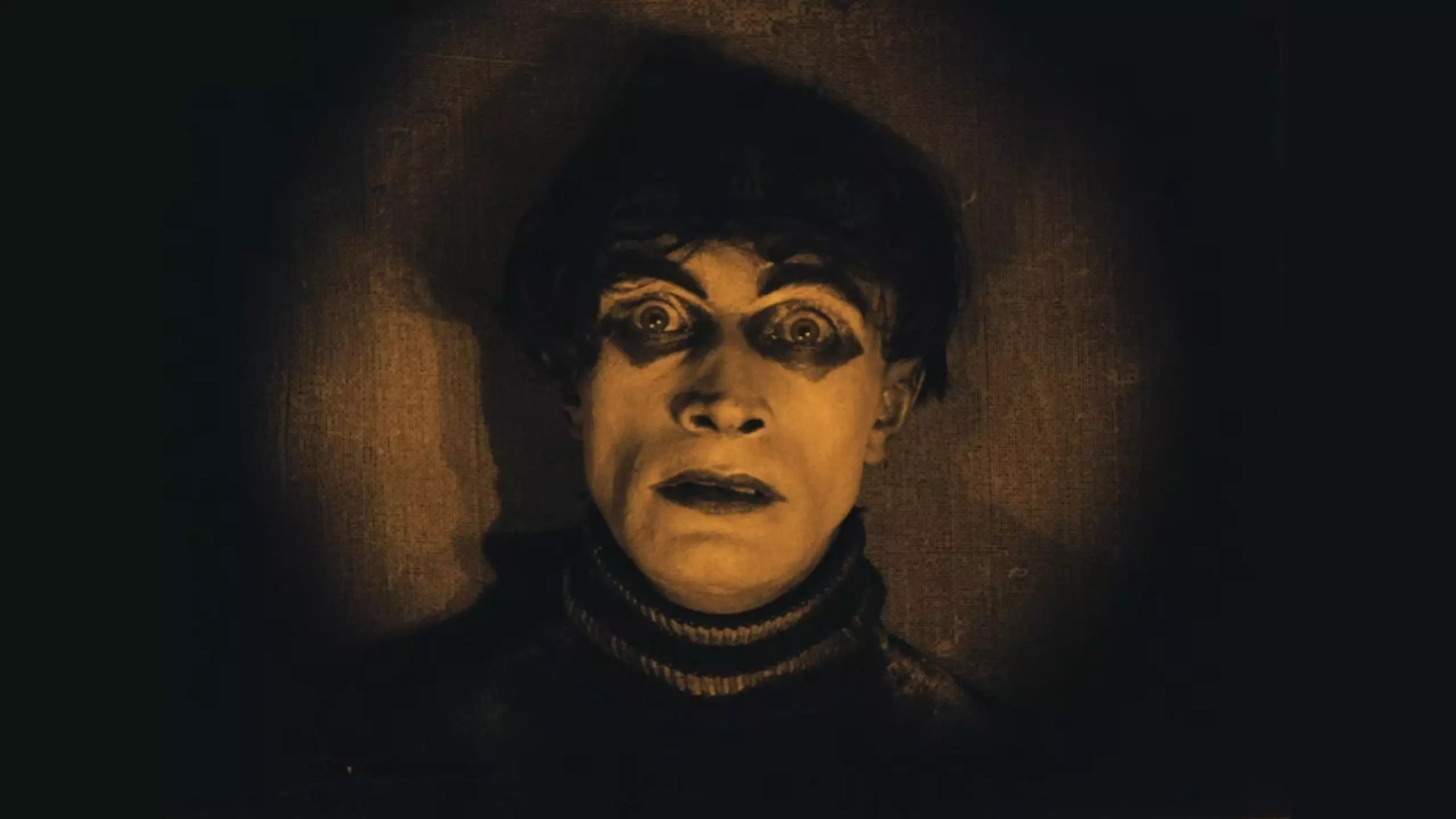 Nahaufnahme eines expressionistisch ausgeleuchteten Männergesichts aus einem Stummfilm, weit geöffnete, geschminkte Augen vor dunklem Hintergrund.