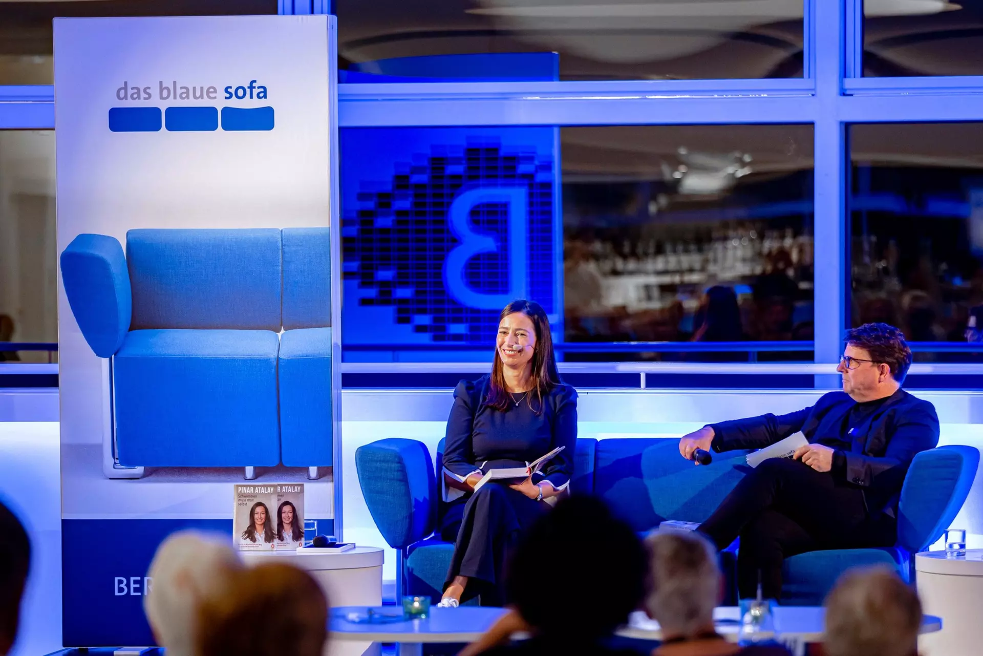 Neben einem Leuchtbanner mit dem Logo des Blauen Sofas sitzen zwei Personen mit Mikrofonen auf einem blauen Sofa. Eine Person hält ein Buch aufgeschlagen in der Hand auf dem Schoß.