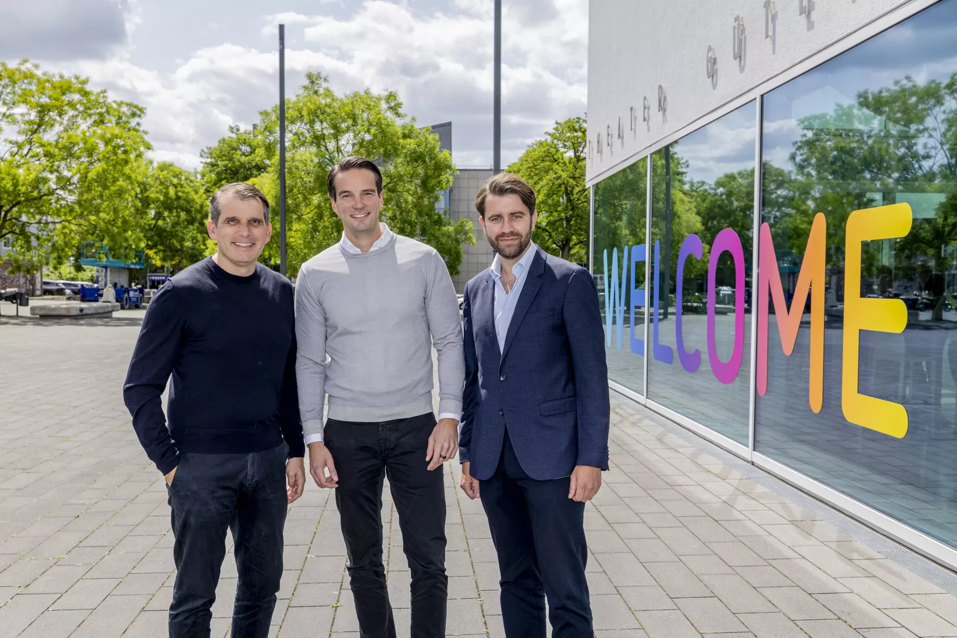 Drei Personen stehen nebeneinander vor Glasfassade mit farbigem Schriftzug „WELCOME“.
