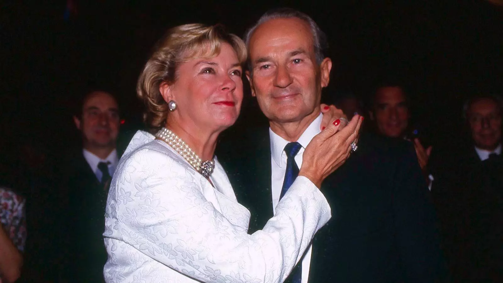 Liz Mohn, in a white suit with a pearl necklace, embraces her husband Reinhard Mohn. They both smile.
