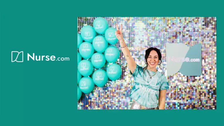 Woman smiles in front of colorful backdrop, raises two fingers, standing beside turquoise balloons with Nurse.com logo.
