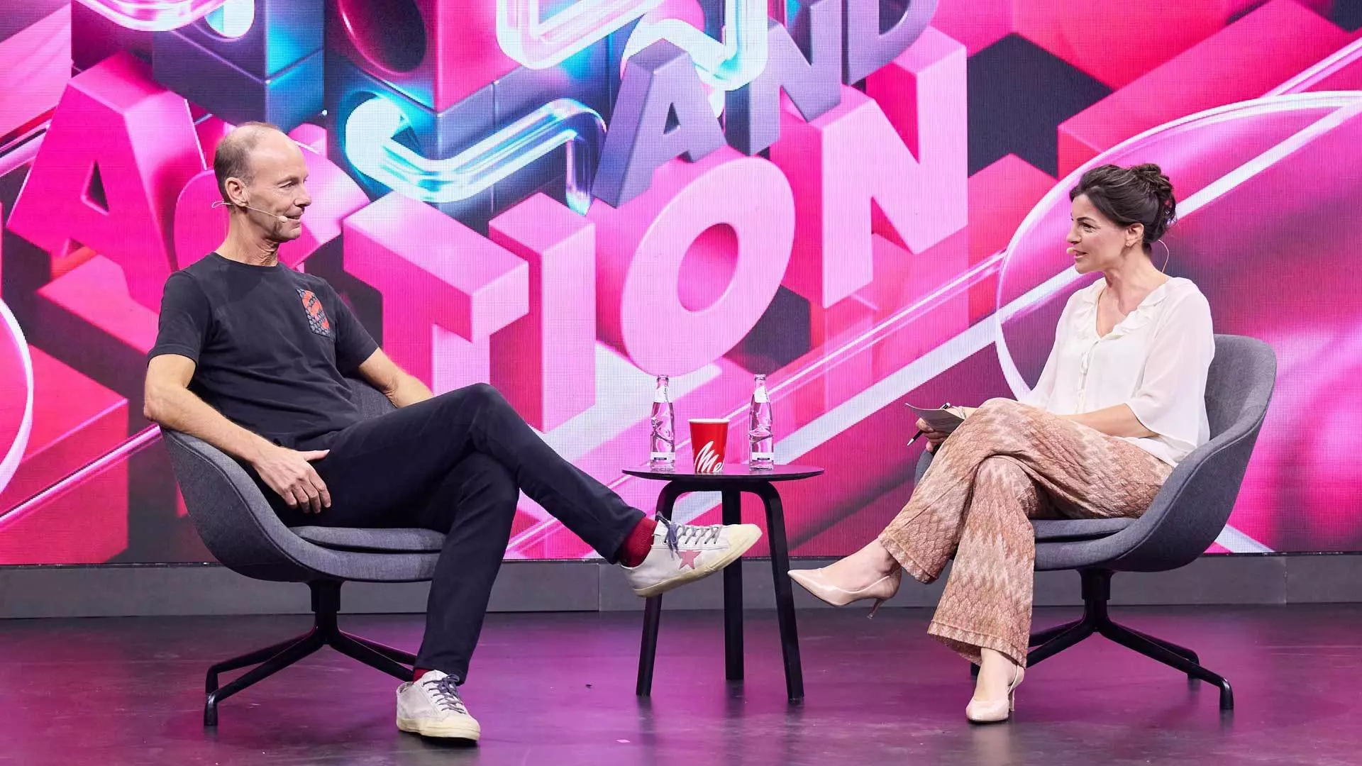 Thomas Rabe and Isabelle Körner are on stage. They are looking at each other. In the middle is a small table with drinks on it.
