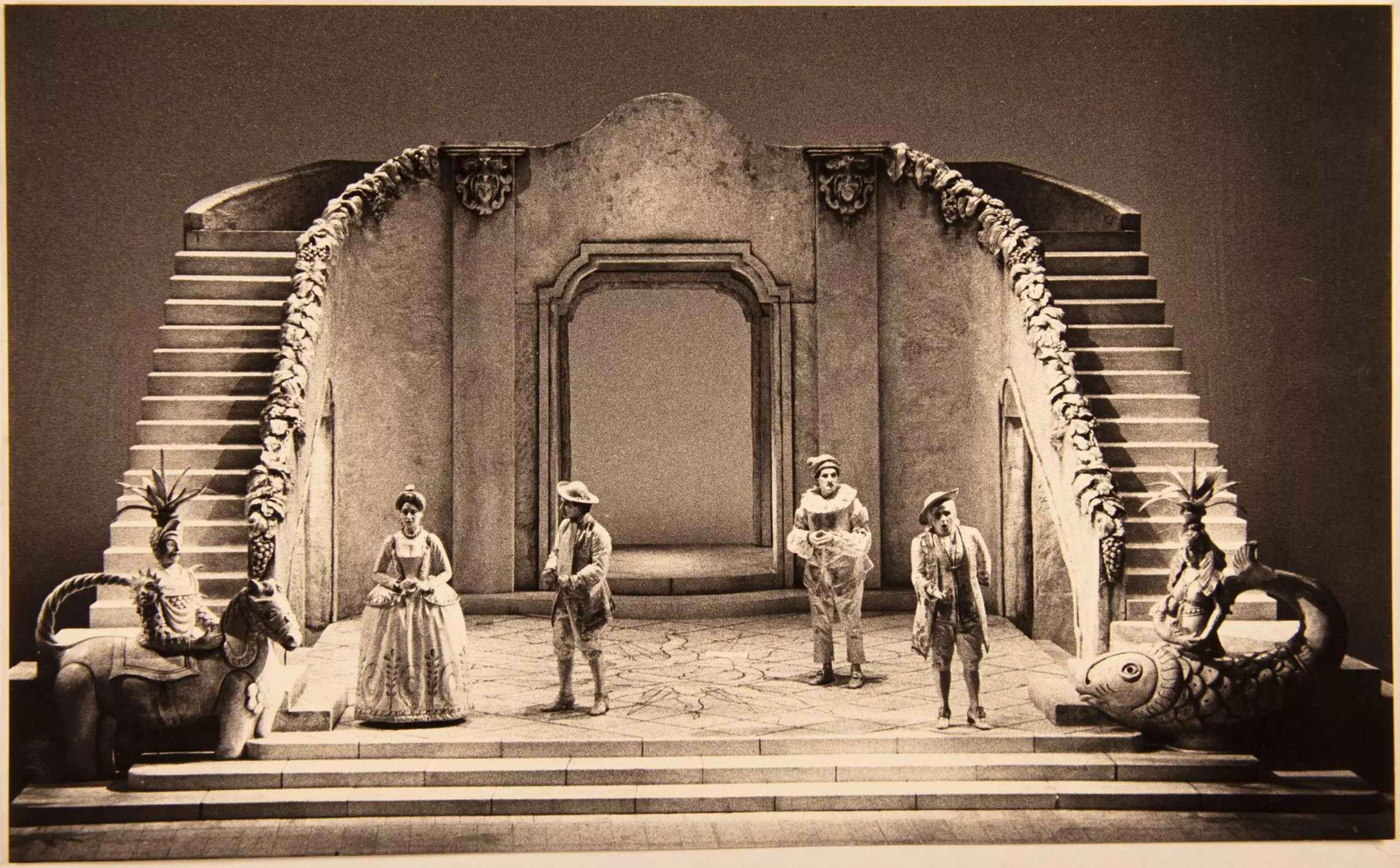 Historische Theaterbühne mit Kulisse aus Treppen und Torbogen, mehrere Darstellende in Kostümen.