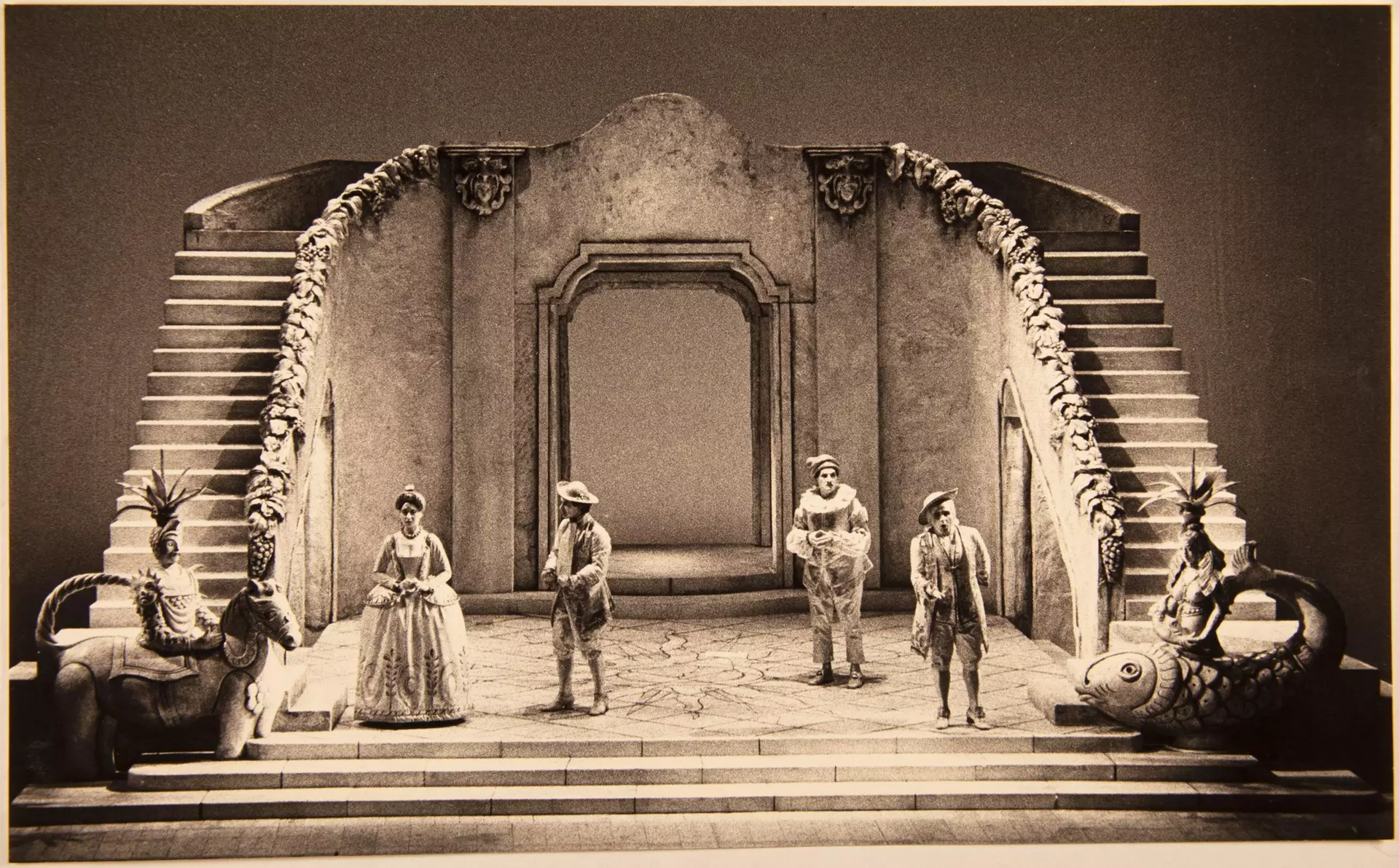 Historische Theaterbühne mit Kulisse aus Treppen und Torbogen, mehrere Darstellende in Kostümen.