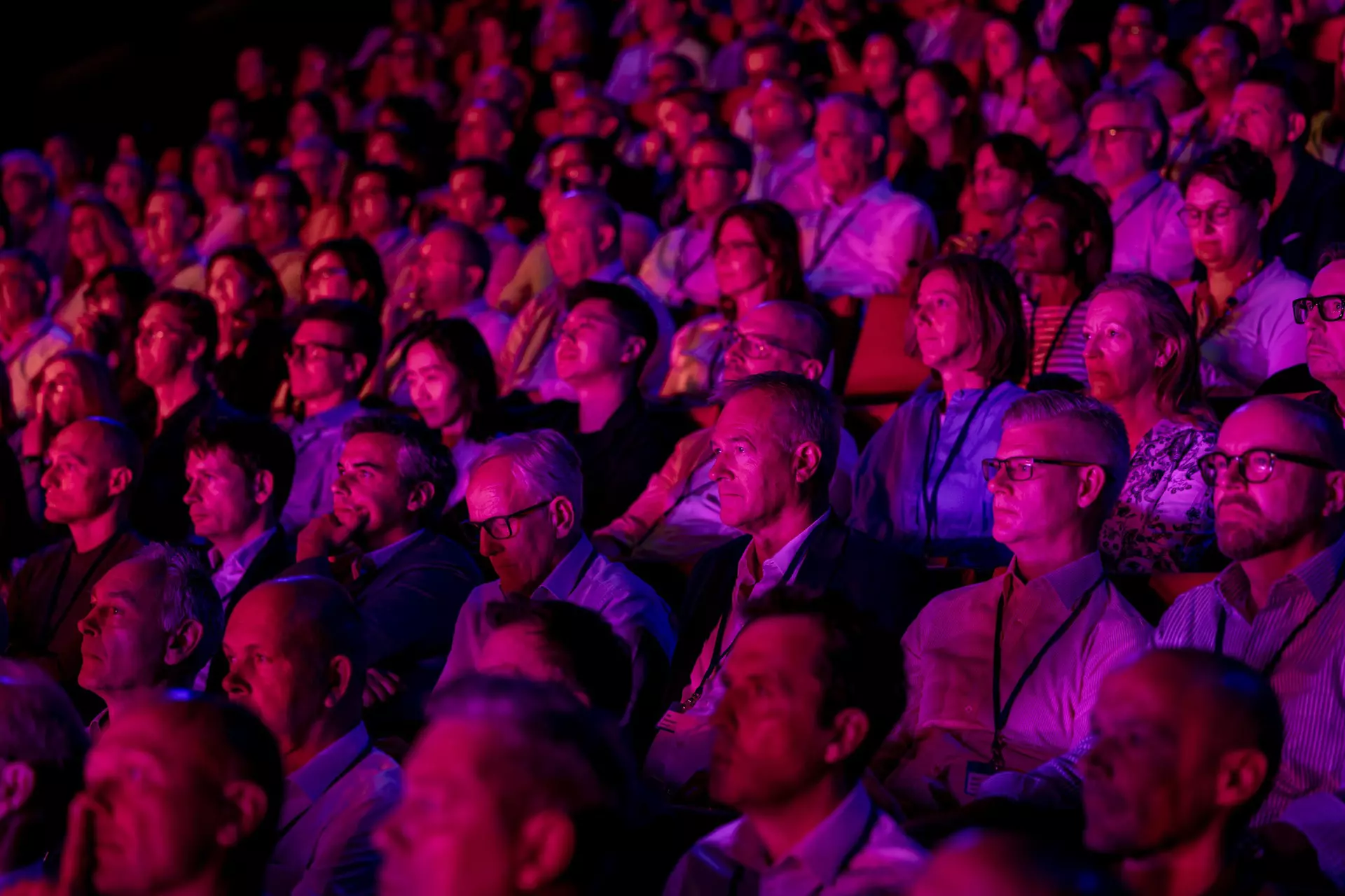 Publikum sitzt dicht gedrängt in Auditorium unter pink-violetter Bühnenbeleuchtung.