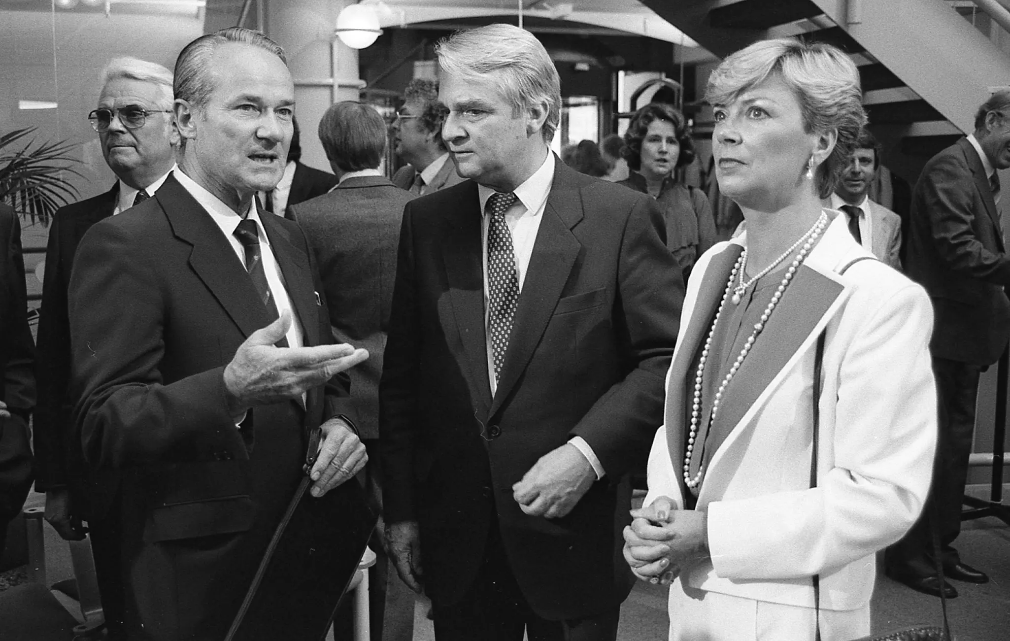 Three people are standing in a large room full of people. Reinhard Mohn is speaking and gesturing with his hand.