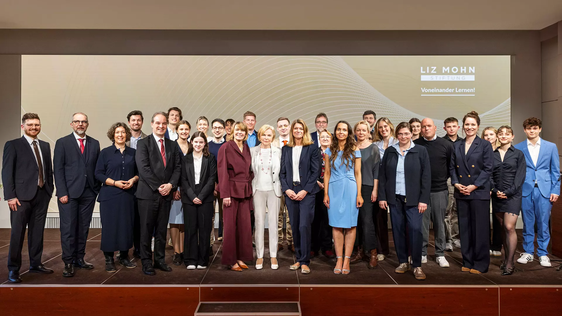 Thirty people are standing on a stage. In the background, the Liz Monh Foundation logo is displayed on a large monitor.