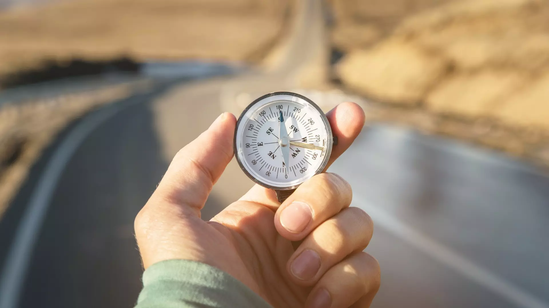 Hand holding a compass on a road in a bright landscape, symbolizing guidance and direction.