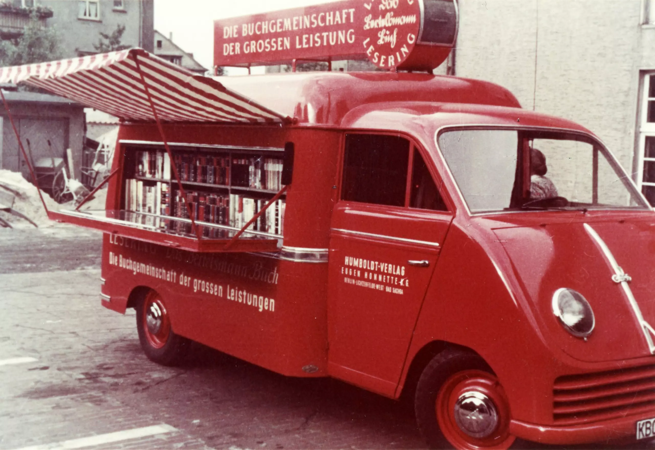 A red, converted camper van with a fold-down counter on the side and a red and white striped canopy containing two rows of books is parked in front of houses. The vehicle bears the inscription: "The Book Club of Great Achievements".