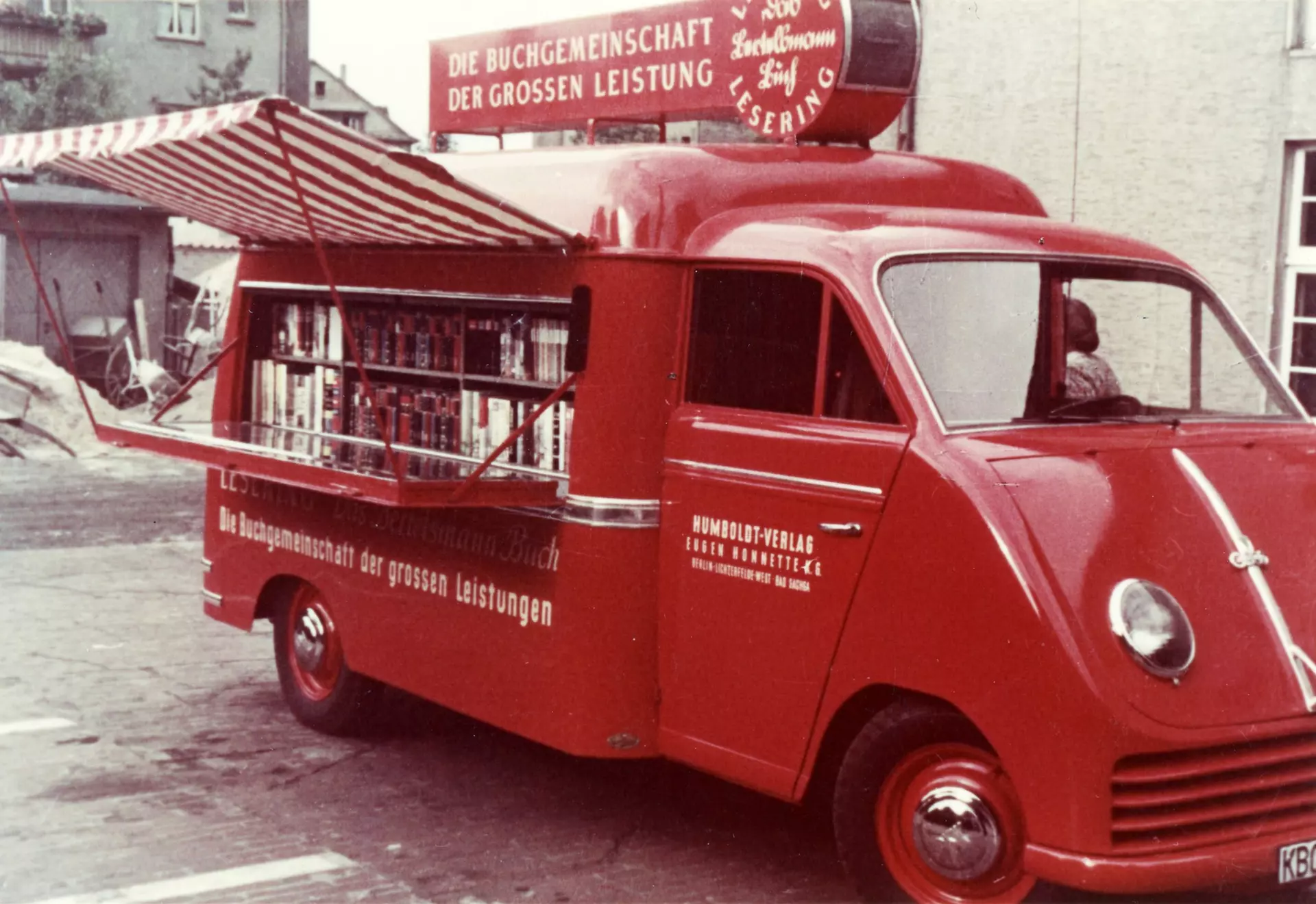 A red, converted camper van with a fold-down counter on the side and a red and white striped canopy containing two rows of books is parked in front of houses. The vehicle bears the inscription: "The Book Club of Great Achievements".