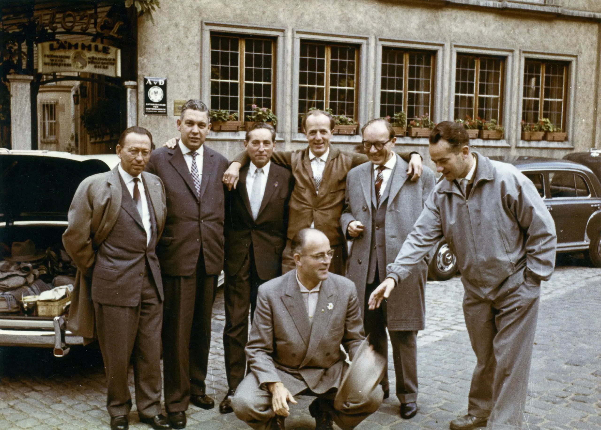A group of five men are standing together, looking cheerful. One man is squatting in front of the group, holding a hat. Reinhard Mohn, standing next to him, extends his hand to him. In the background is a restaurant with five casement windows.