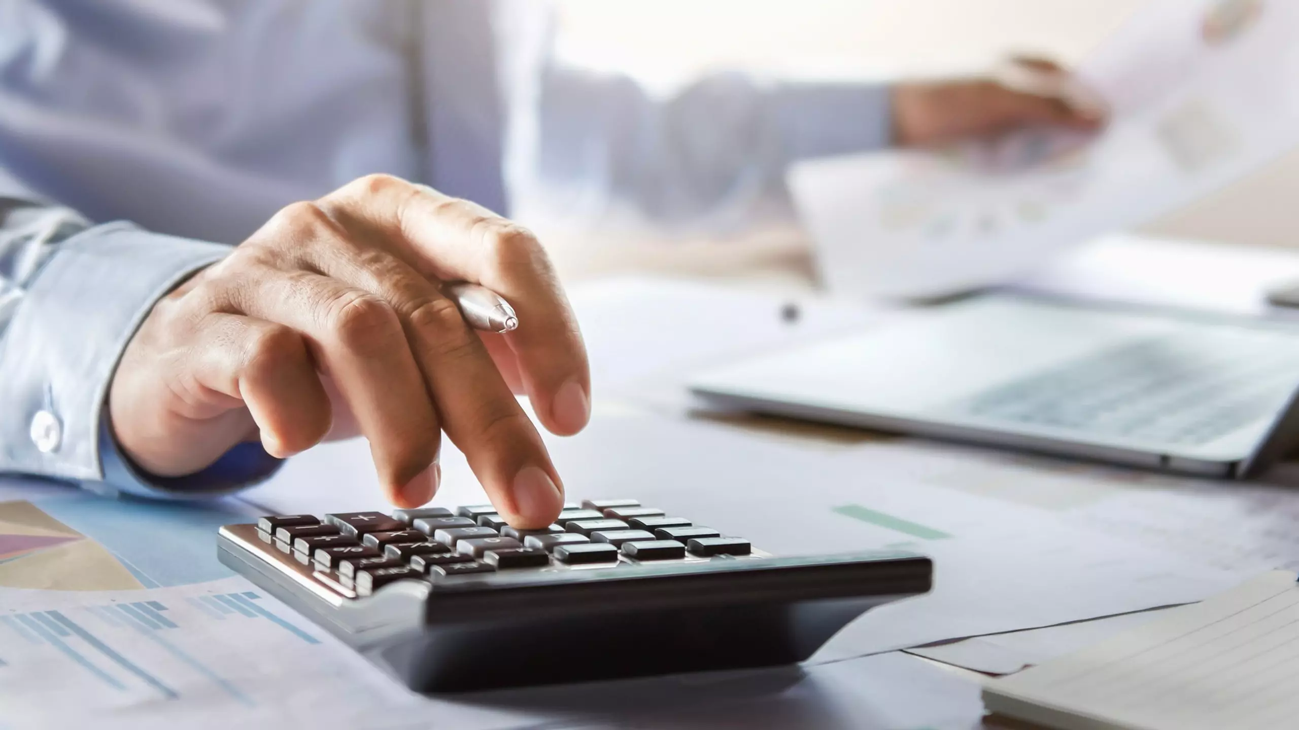 Hand using a calculator, with documents and a laptop nearby.