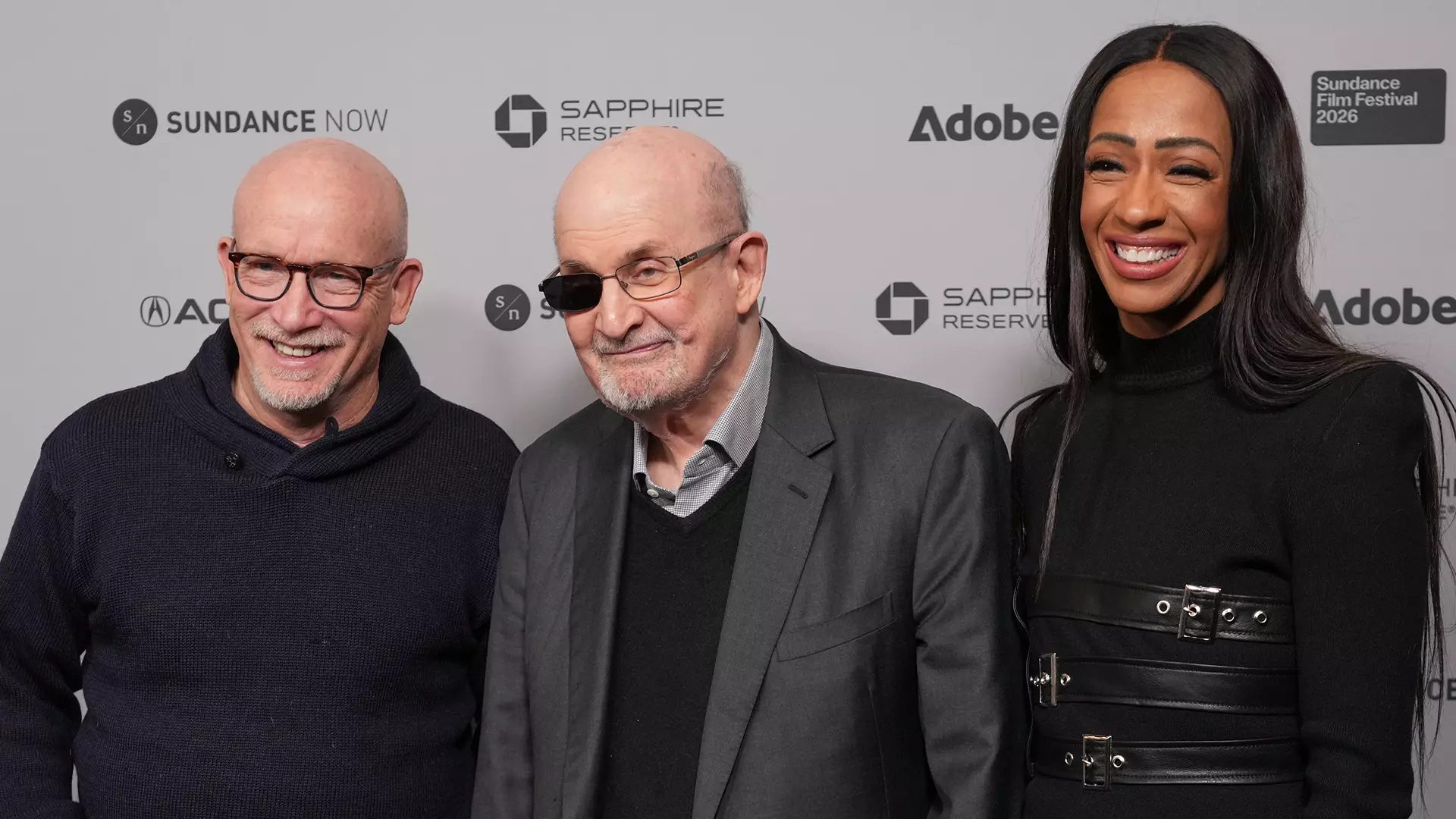 Two men stand in front of the Sundance Film Festival logo wall. On the left is Alex Gibney, a bald man wearing horn-rimmed glasses and a black sweater; on the right is Salman Rushdie wearing glasses that darken his right eye. He has little hair and wears a gray jacket over his plaid shirt. Next to him stands his tall, dark-skinned wife, Eliza Griffiths, dressed in black. They are all smiling at the camera.
