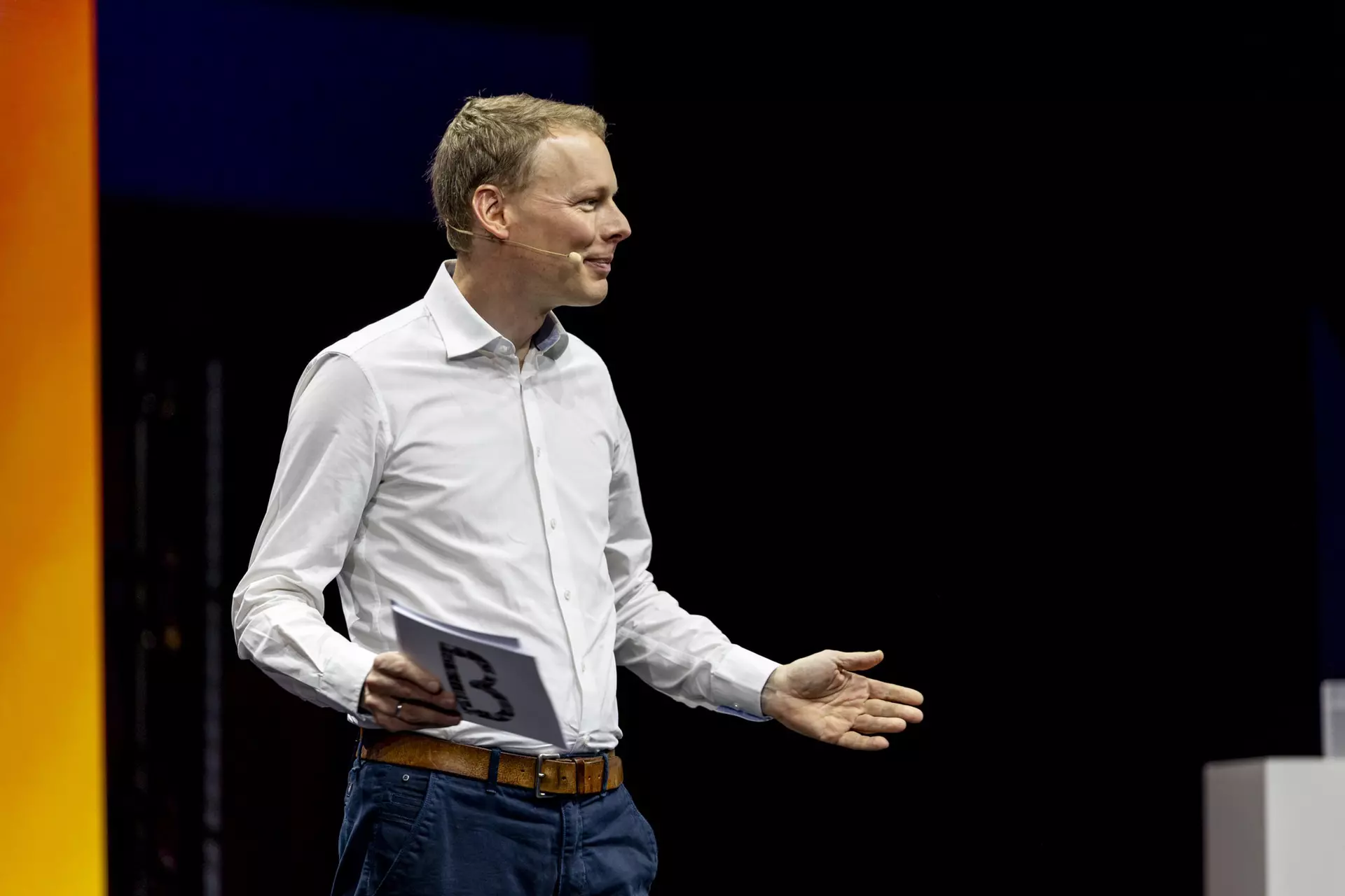 Person im weißen Hemd mit Karte in der Hand spricht auf Bühne vor dunklem Hintergrund.