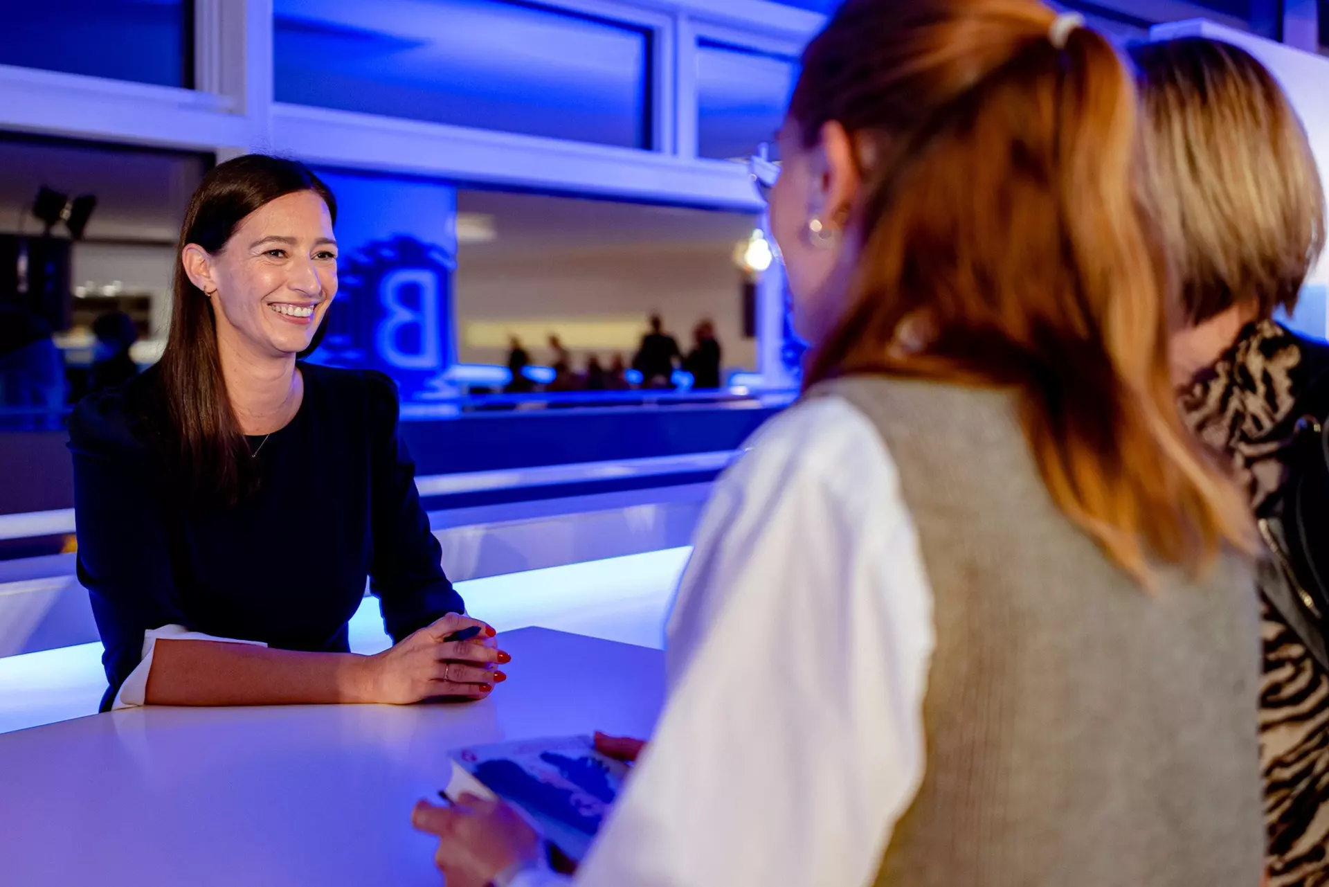 Die Autorin an einem Stehtisch schaut lächelnd zwei Gäste an während des Signierens ihres Buches. Im Hintergrund spiegelt sich in der Fensterfront die Skylobby des Theaters Gütersloh mit weiteren Gästen.