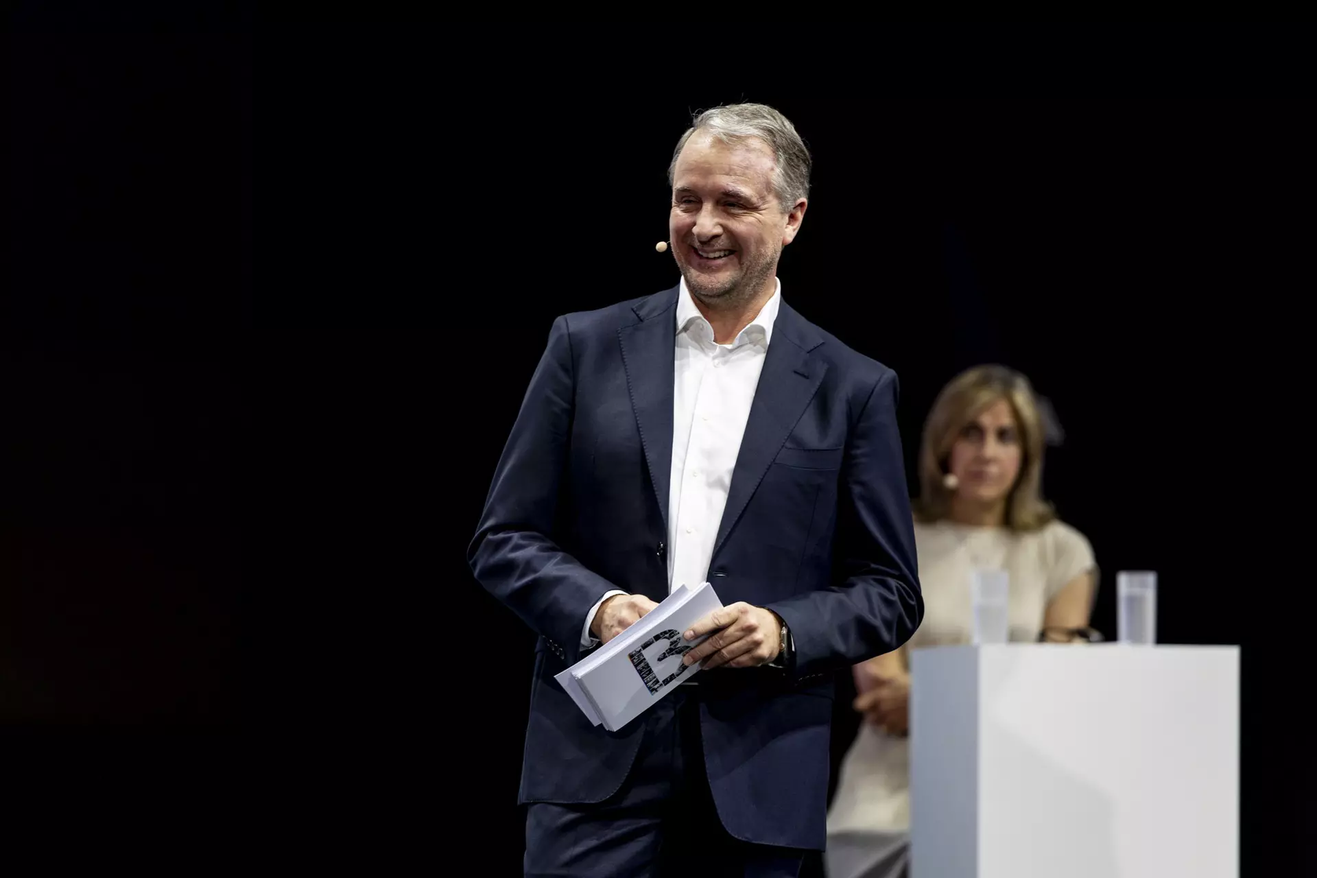Person im dunklen Anzug mit Unterlagen auf Bühne, im Hintergrund Person an Rednerpult.