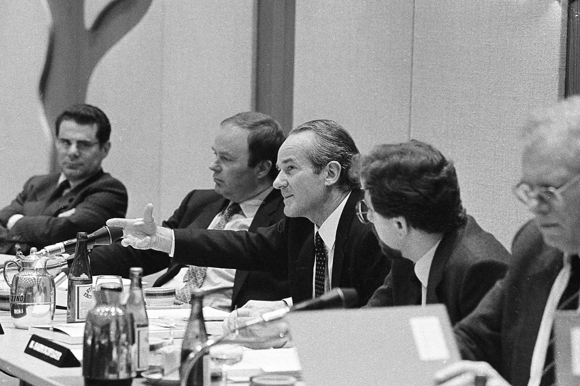 Five people are sitting at a long table with table microphones pointed at them. Reinhard Mohn sits in the middle and gestures with his right hand.