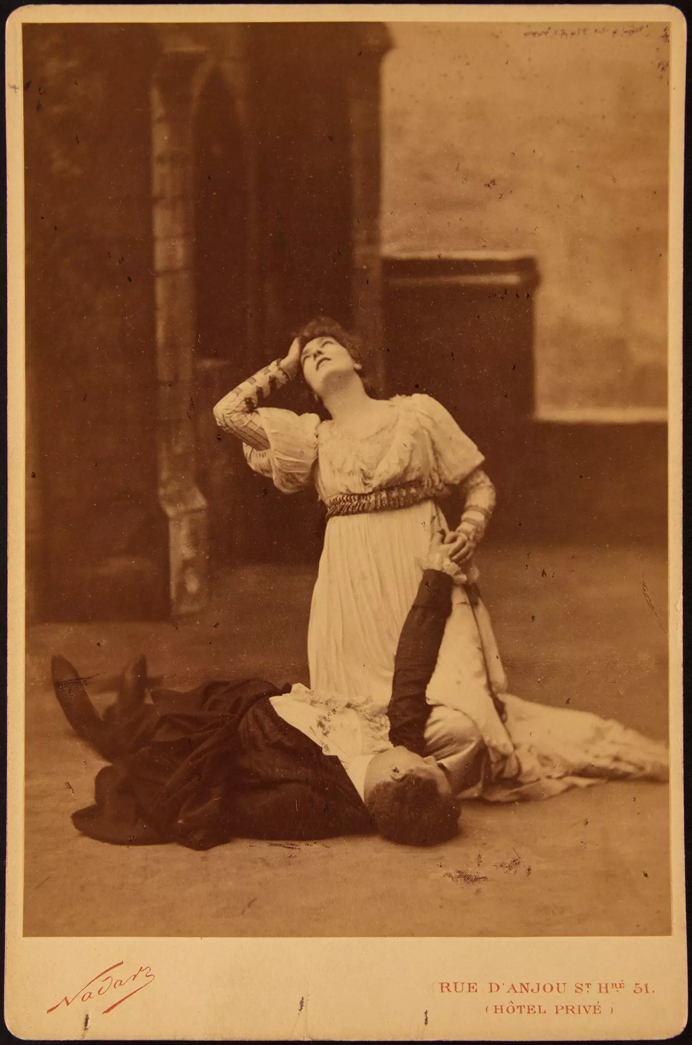 Historisches Bühnenfoto mit Frau im hellen Gewand über liegender Person, dramatische Szene vor Kulisse.