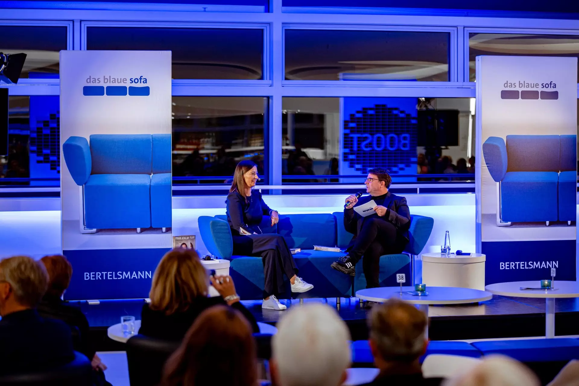 Die Autorin und der Moderator sitzen auf dem blauen Sofa und unterhalten sich. Im Vordergrund sind die Hinterköpfe von Zuschauern zu sehen.