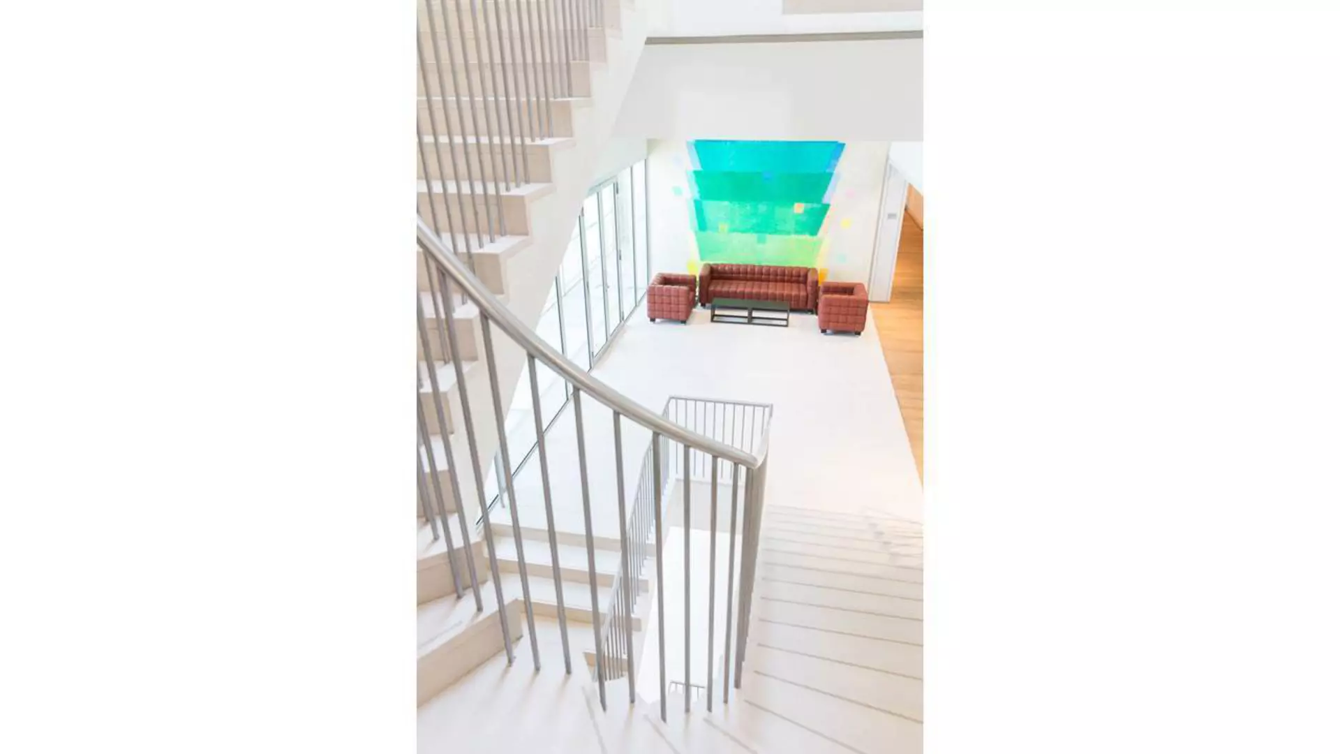 Treppenhaus mit geschwungener Brüstung, Blick in helles Foyer mit Sitzgruppe vor farbigem Wandkunstwerk.