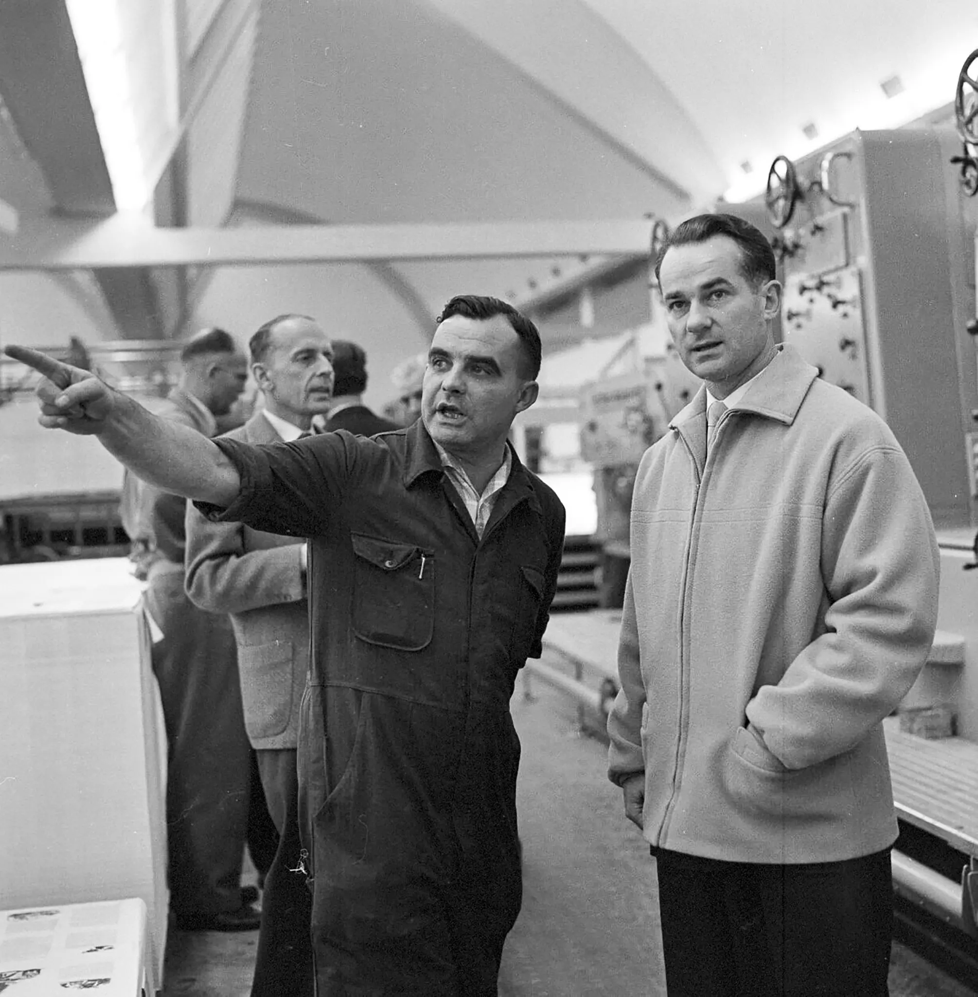 An employee in a work suit stands in front of machines and points to something in the distance while Reinhard Mohn looks in the same direction.