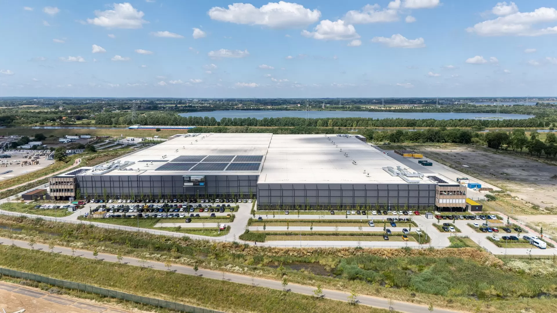 An aerial view of a large white logistics center, with cars in front of it and a lake behind it.
