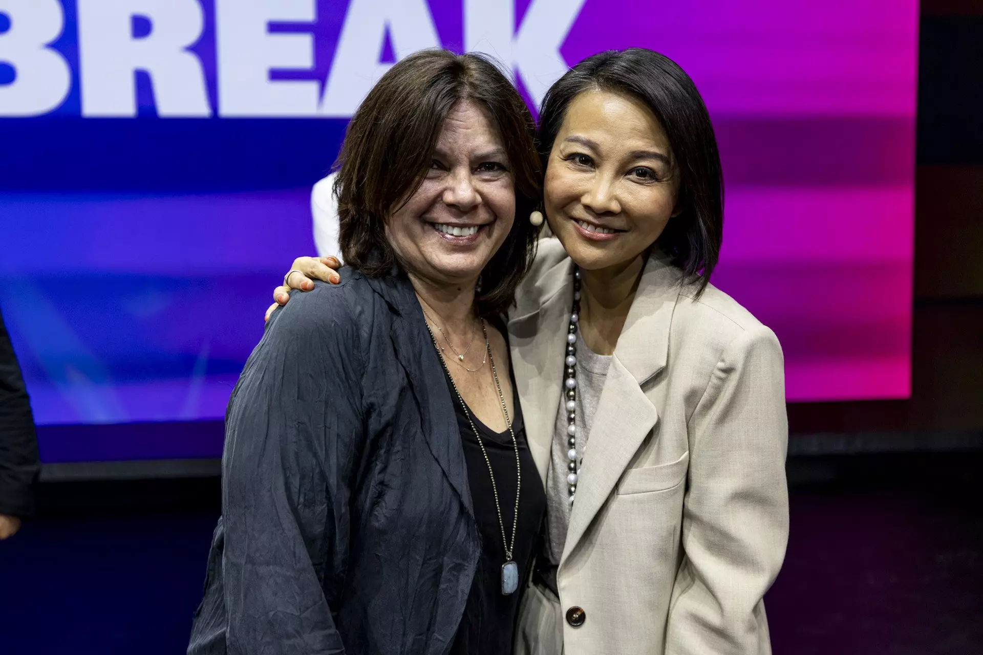 Zwei Personen umarmen sich lächelnd auf Bühne vor pink-blauem Hintergrund.