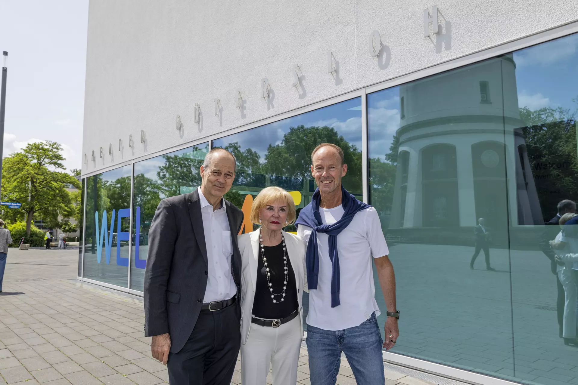 Drei Personen stehen vor Glasfassade mit Schriftzug „WELCOME“ und spiegelnder Gebäudearchitektur.