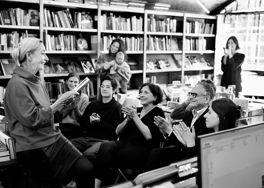 Person liest aus Buch vor sitzendem, applaudierendem Publikum, im Hintergrund Regale voller Bücher.