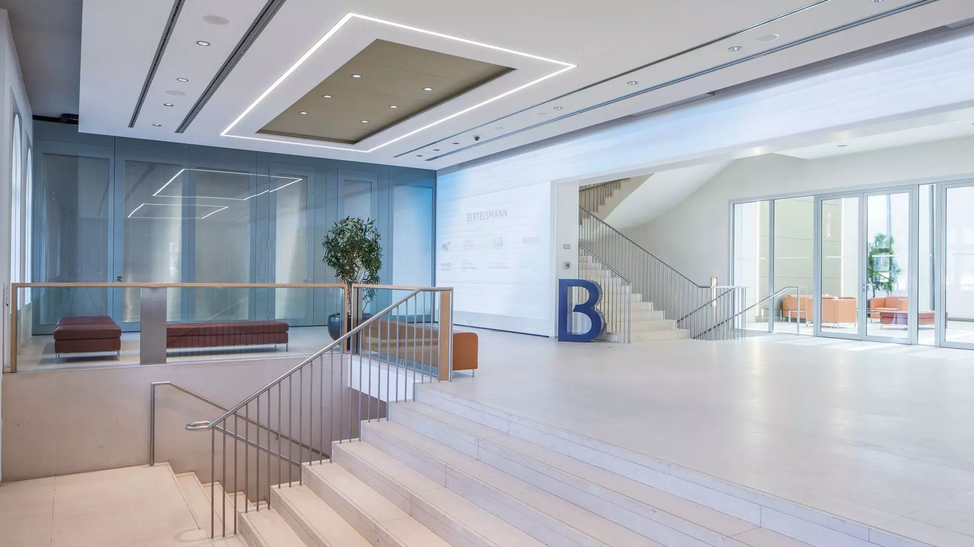Großzügiges Foyer mit breiter Treppe, Glasgeländern, Sitzbereichen, Wandgrafik und abgehängter Decke mit Licht.