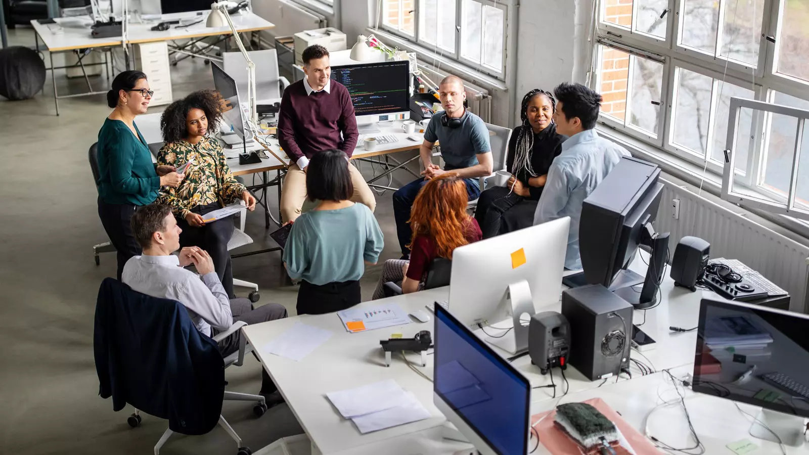 Nine people are standing in a group in the office, engaged in lively conversation.