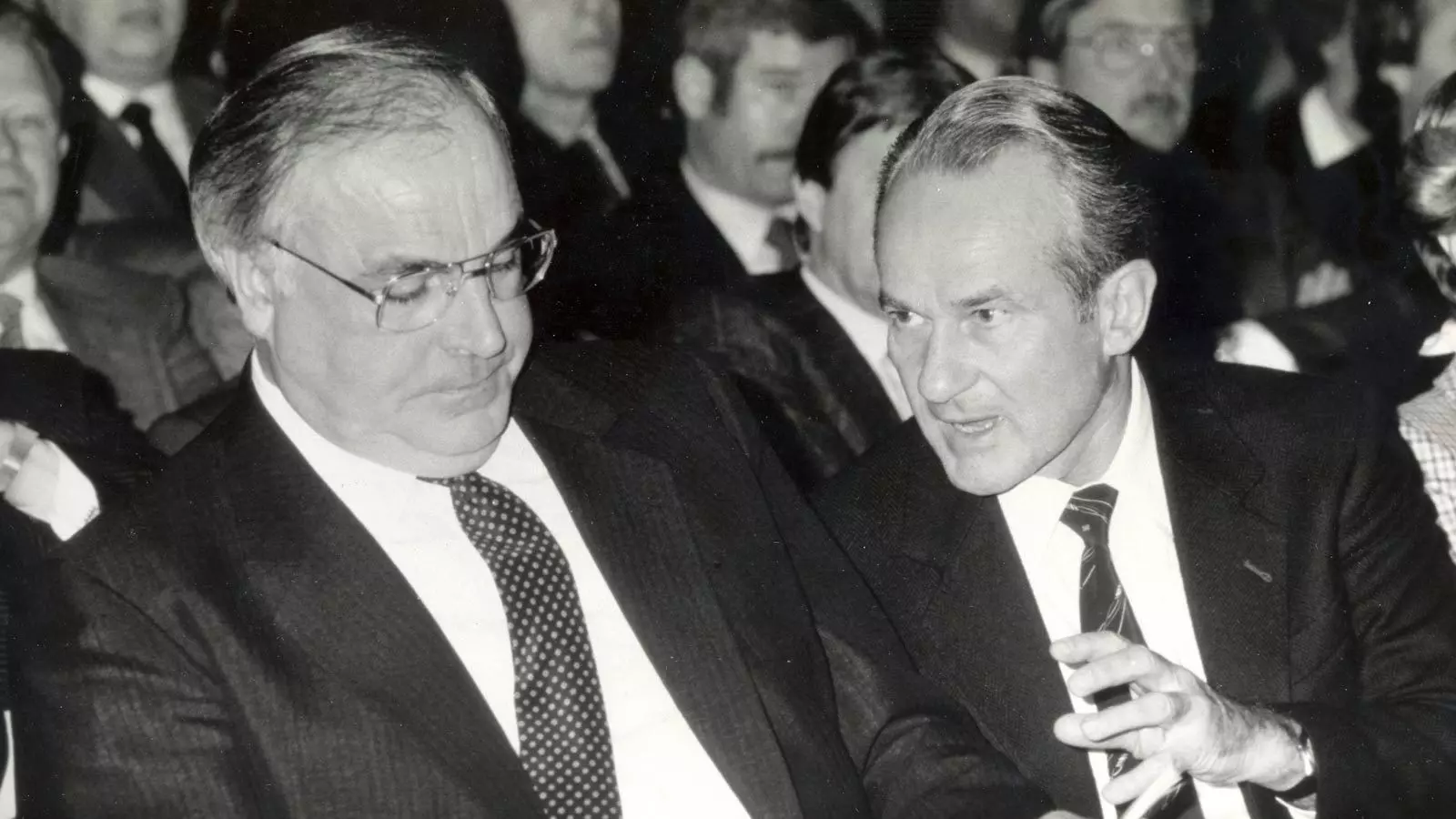 Helmut Kohl is sitting next to Reinhard Mohn, who is talking to him. They are sitting in a room with many people sitting in rows of chairs behind them.