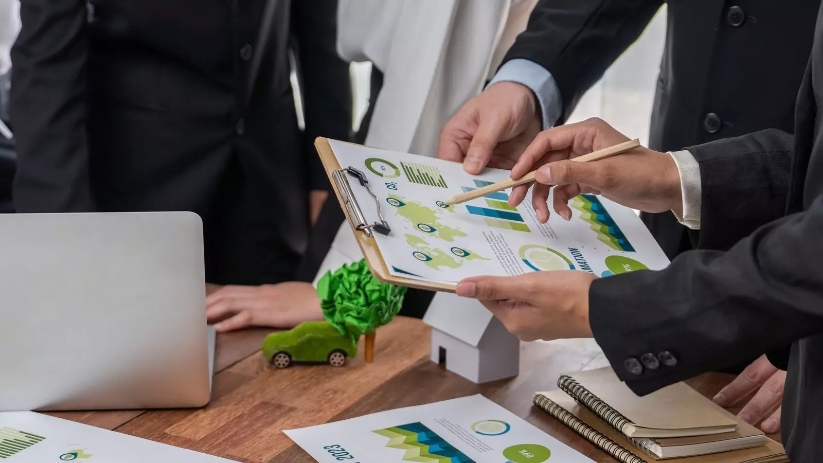 Three people working on sustainability charts on a clipboard; laptop and office supplies on the table.