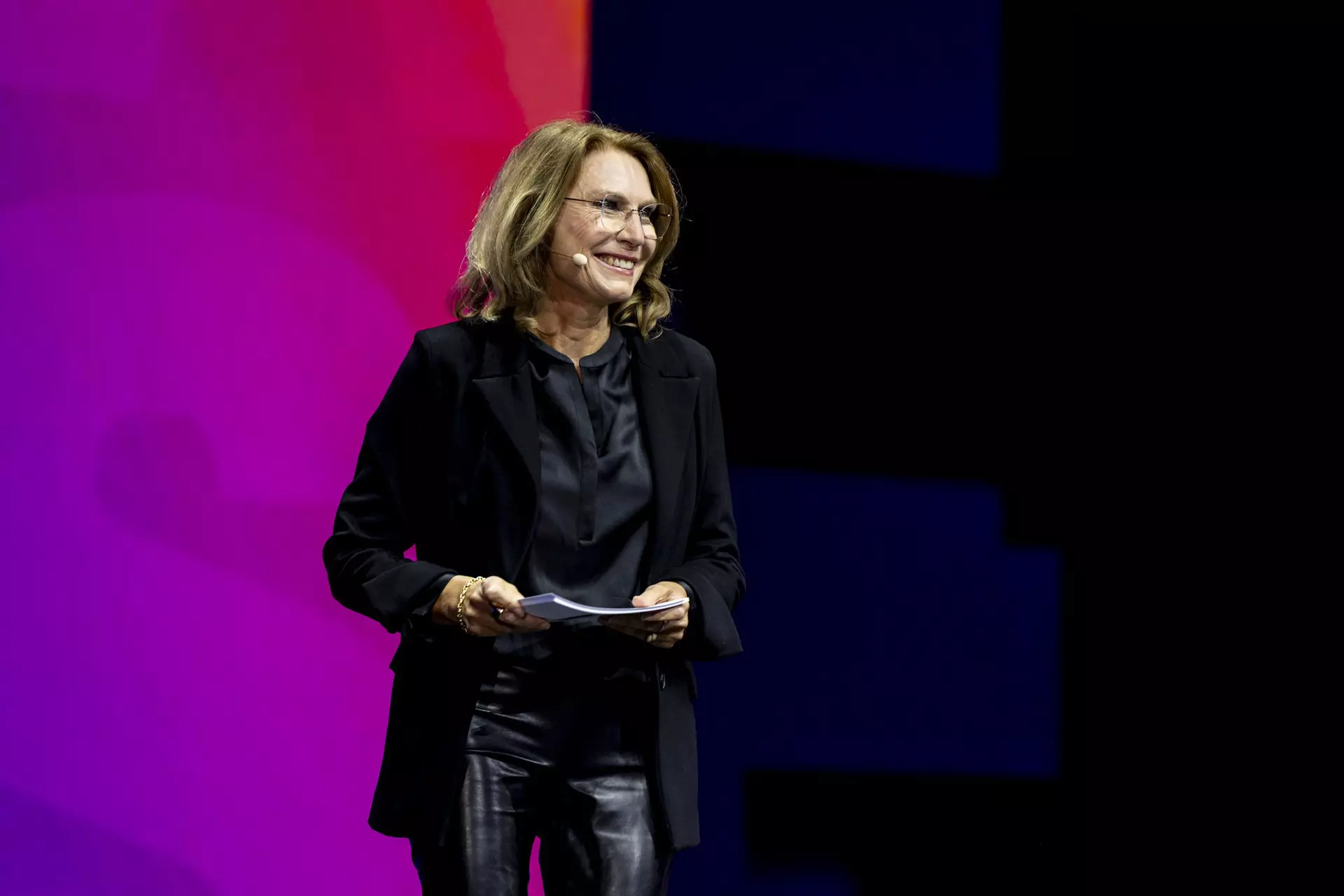 Person in schwarzer Kleidung steht auf Bühne vor pink-blauem Hintergrund