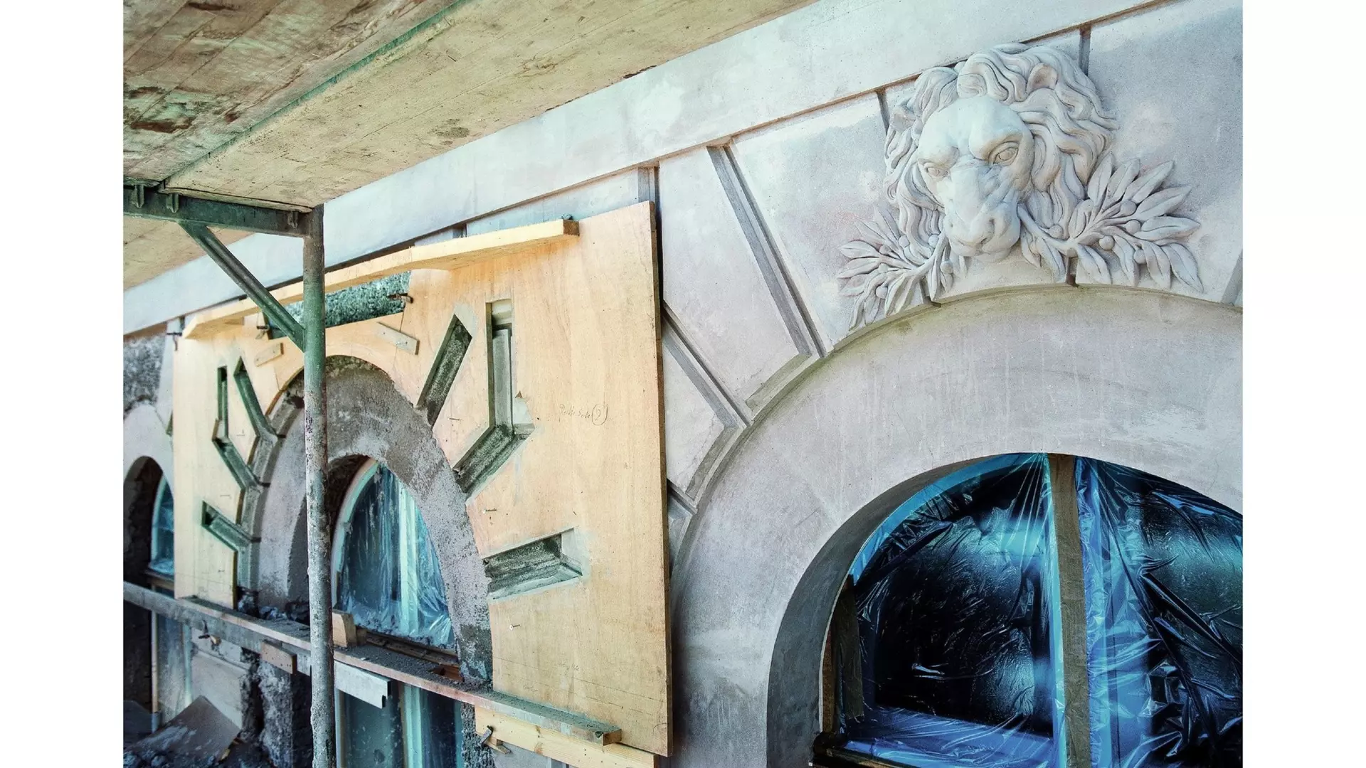 Fassadenbereich mit Gerüst, abgedeckten Rundbogenfenstern und Löwenrelief über einem Bogen während Bauarbeiten.